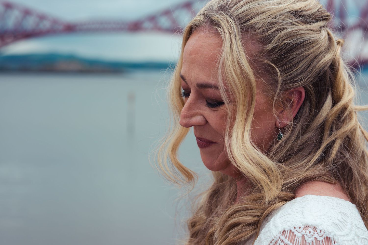 Blonde woman in white lace dress gazes down near Forth Bridge, Scotland, with river in background.