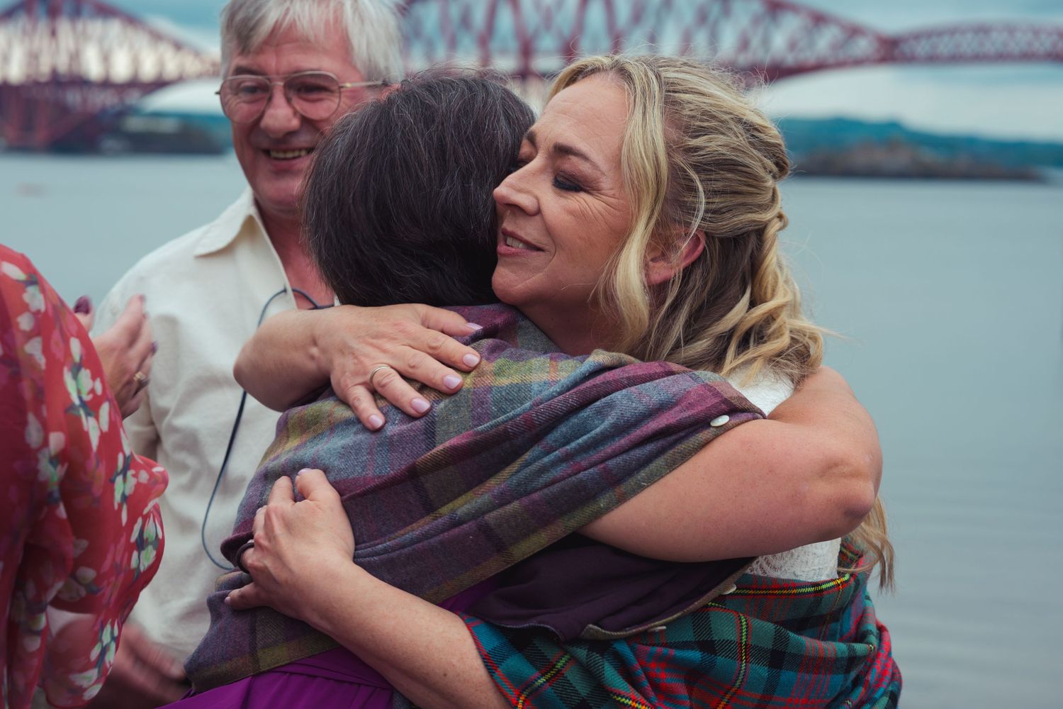 People sharing a warm group hug outdoors near a bridge, smiling joyfully on a cloudy day.