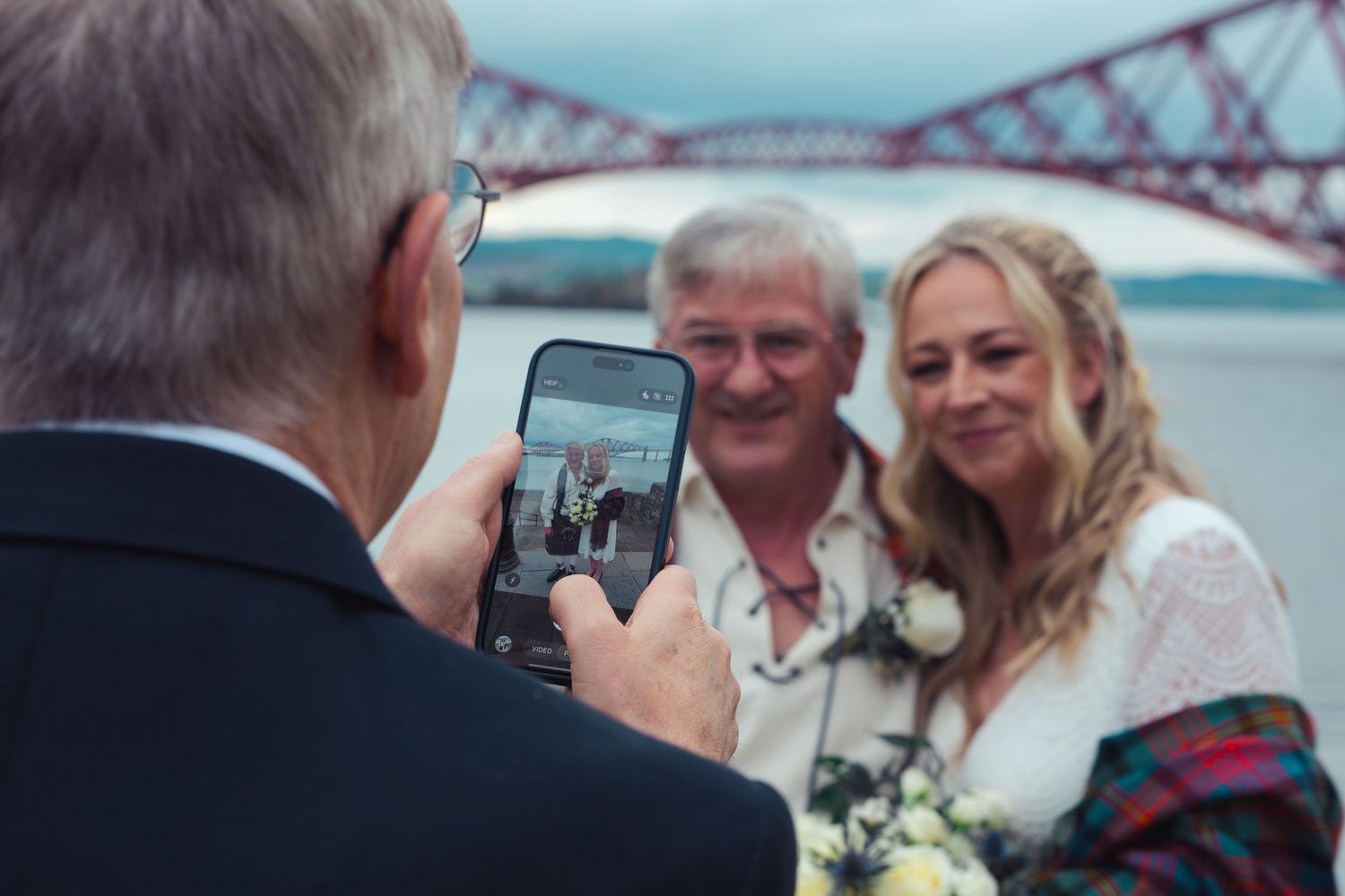 Man photographing smiling bride and groom with Forth Rail Bridge in background at Scottish wedding.