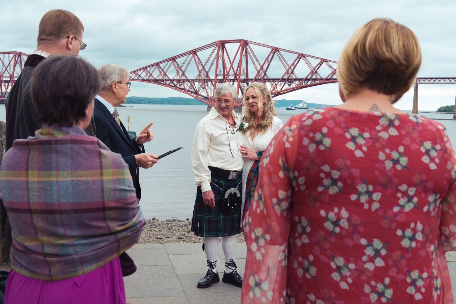 Outdoor wedding ceremony near the Forth Bridge in Scotland, with groom in kilt and bride among guests.