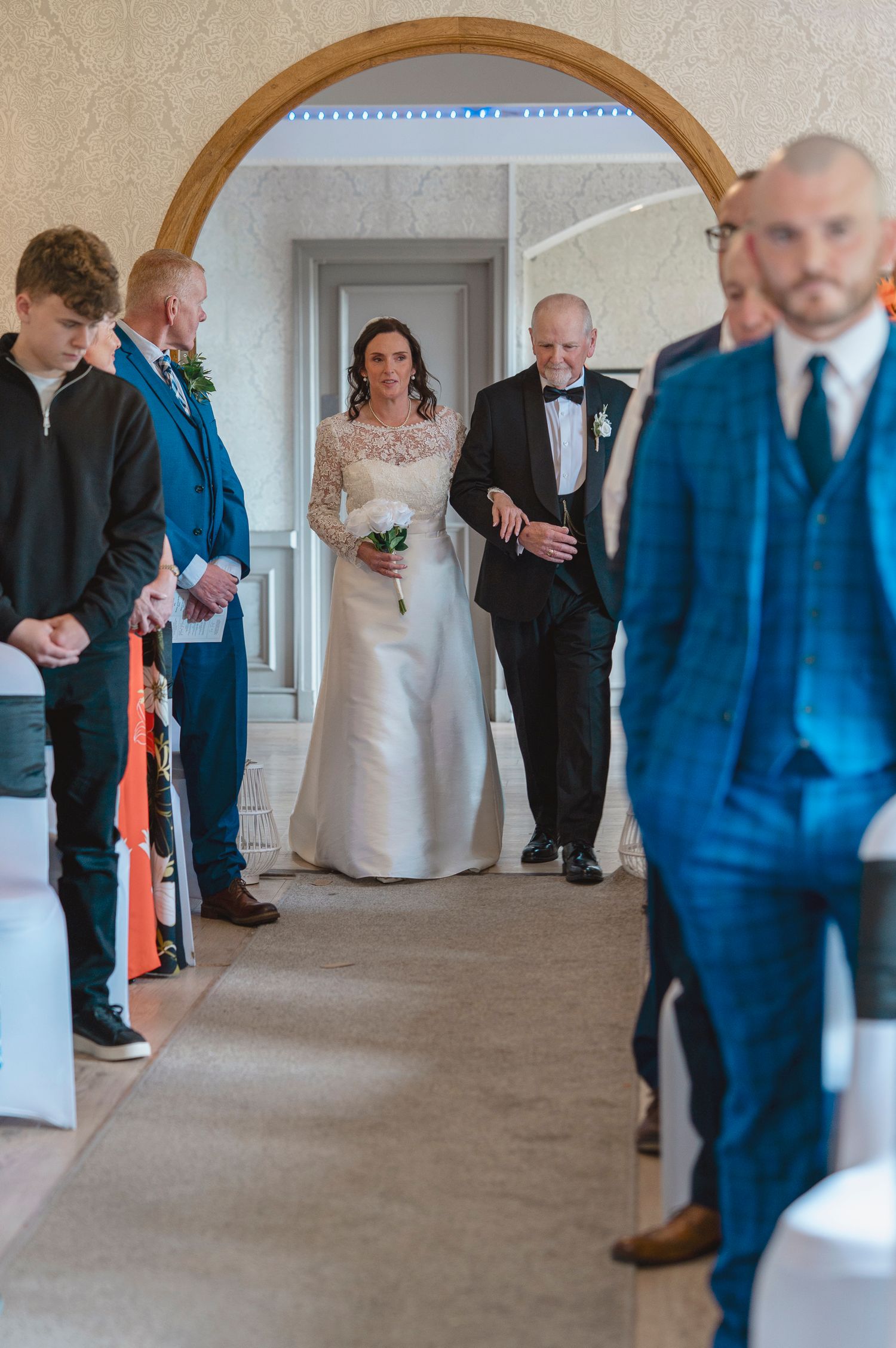 Bride walks down the aisle with escort through arch doorway as guests and groom await in elegant wedding venue.