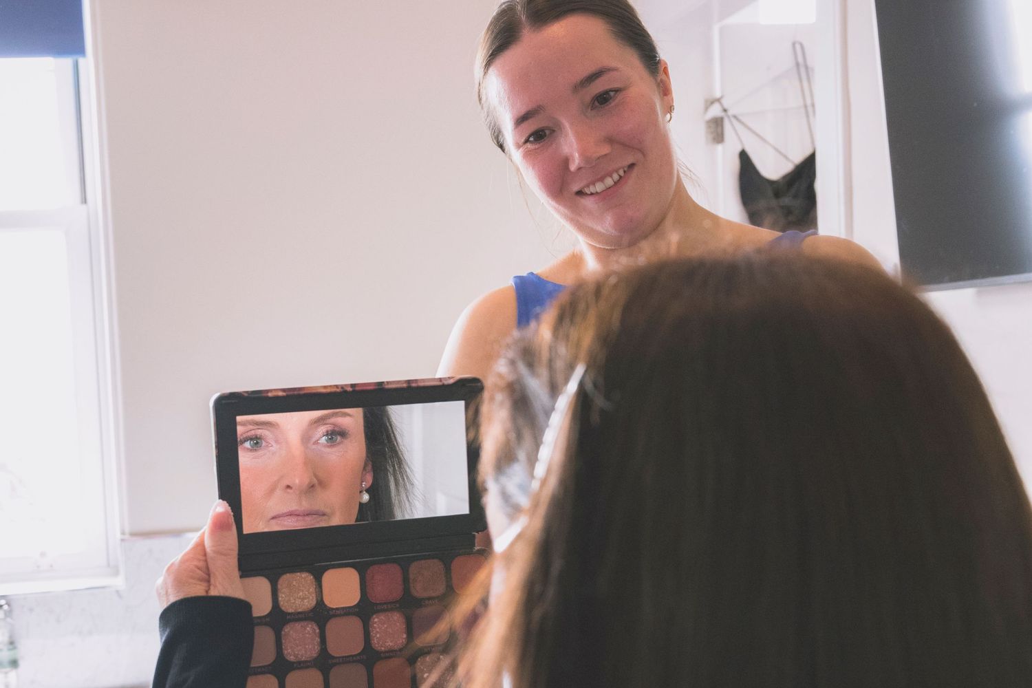 A makeup artist holds a palette mirror showing a woman's reflection while applying makeup in a bright room.