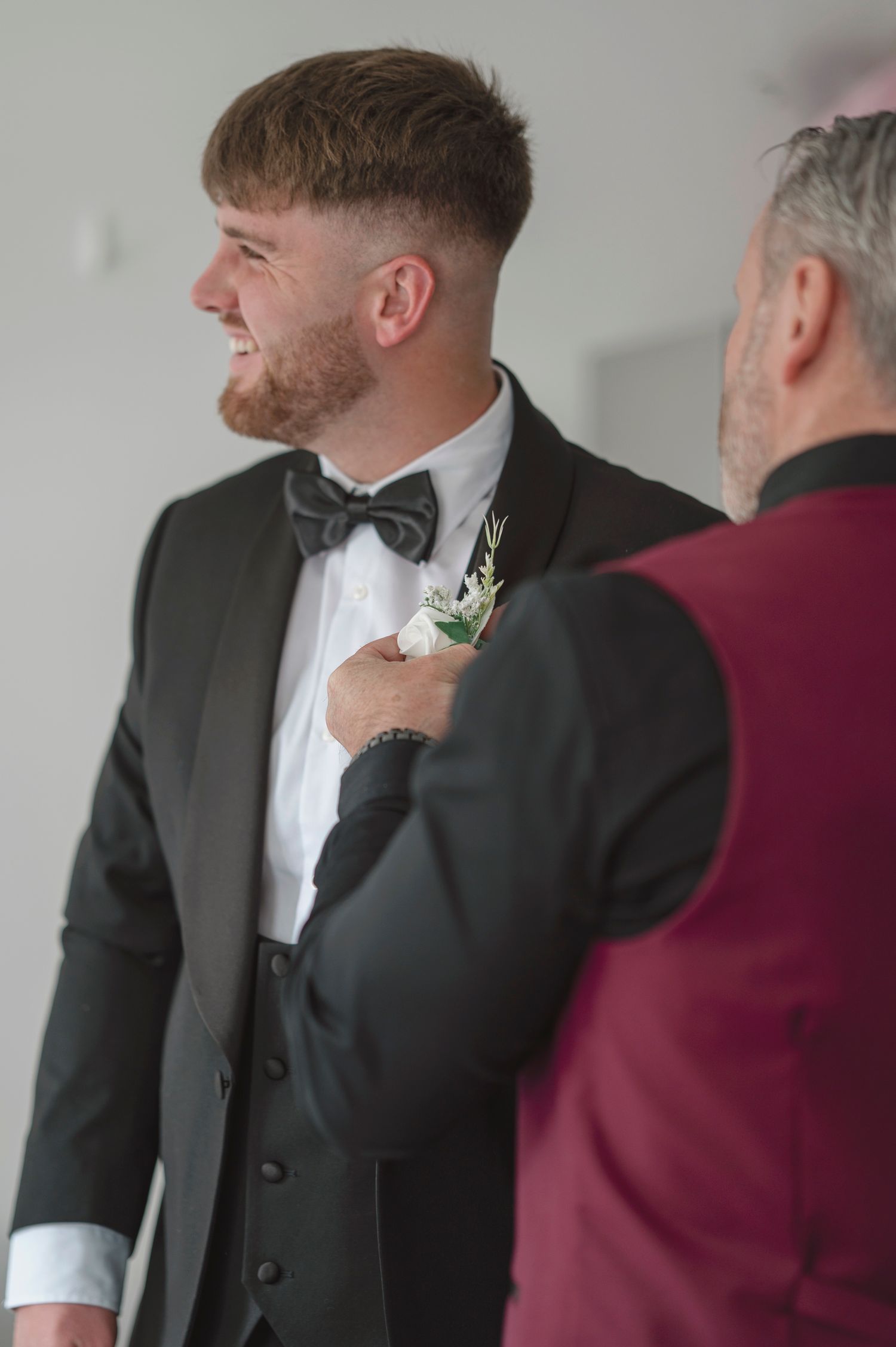 Groom in black tuxedo and bow tie smiles as man in burgundy jacket pins boutonniere on his lapel.