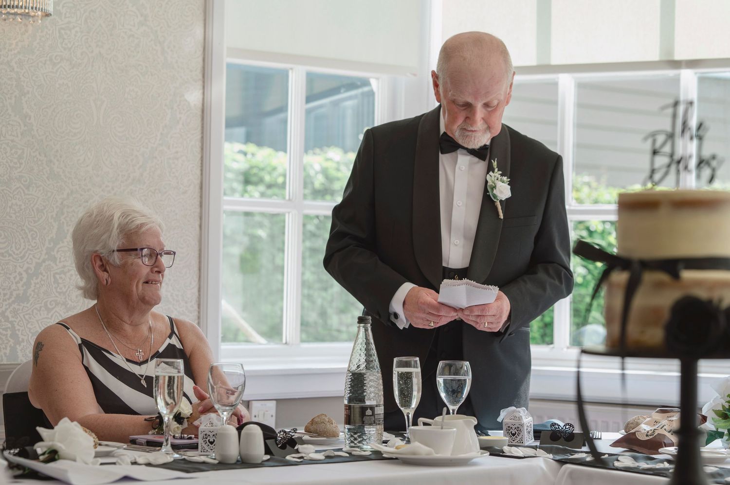 Elderly man in tuxedo giving wedding speech while woman in black and white dress sits at decorated reception table.