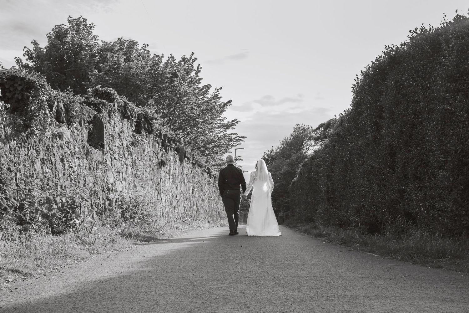 Black and white photo of a bride and groom walking hand in hand down a quiet country road lined with stone walls.