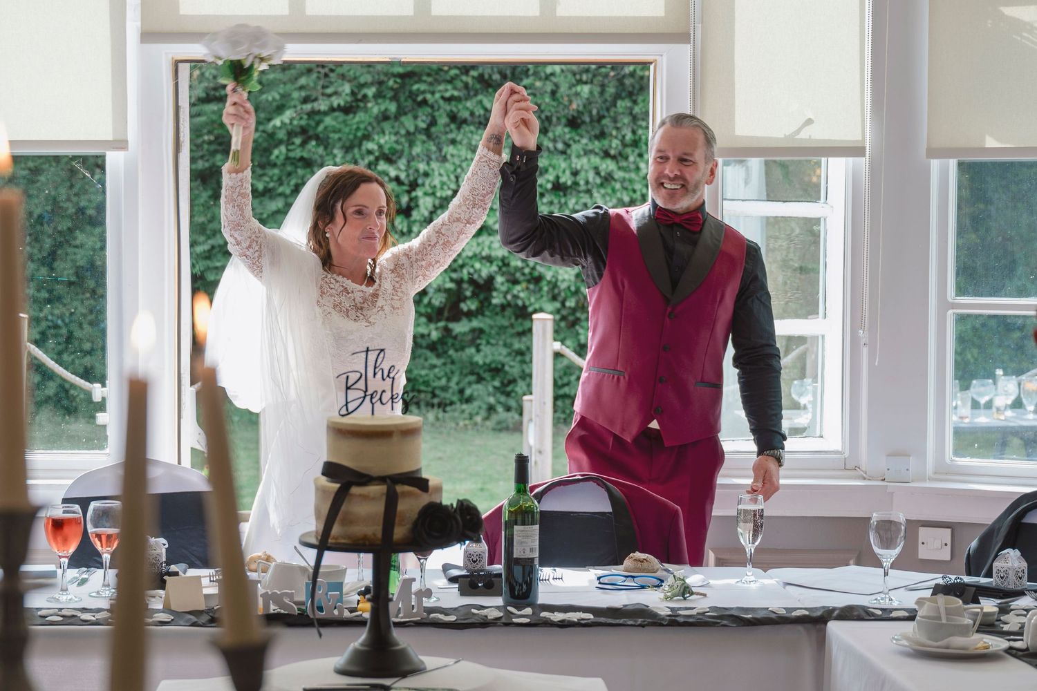 Bride and groom celebrate at wedding reception, raising hands in joy near decorated table and window.