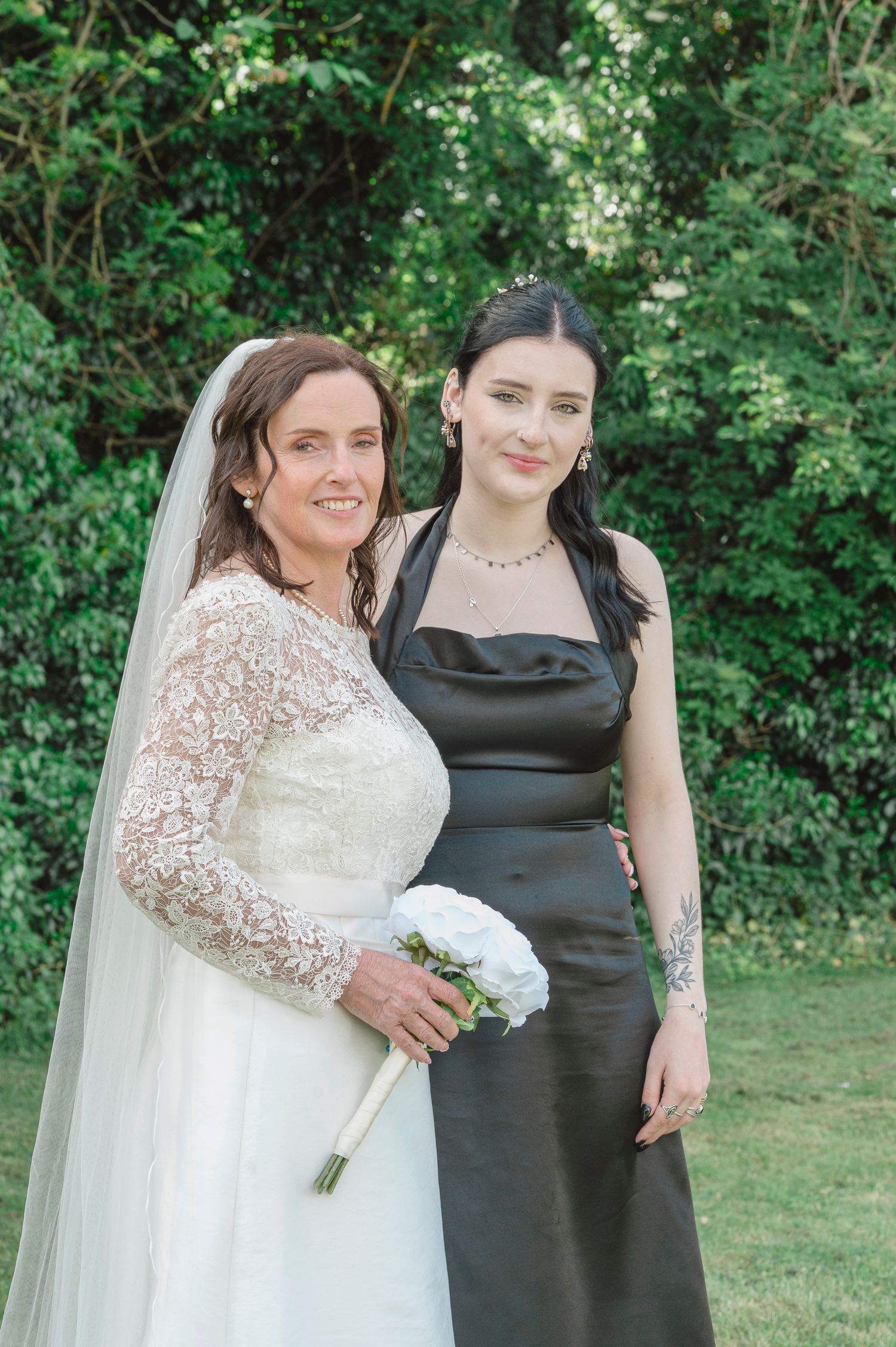Bride in white lace gown with veil holding bouquet stands with bridesmaid in black satin dress outdoors.