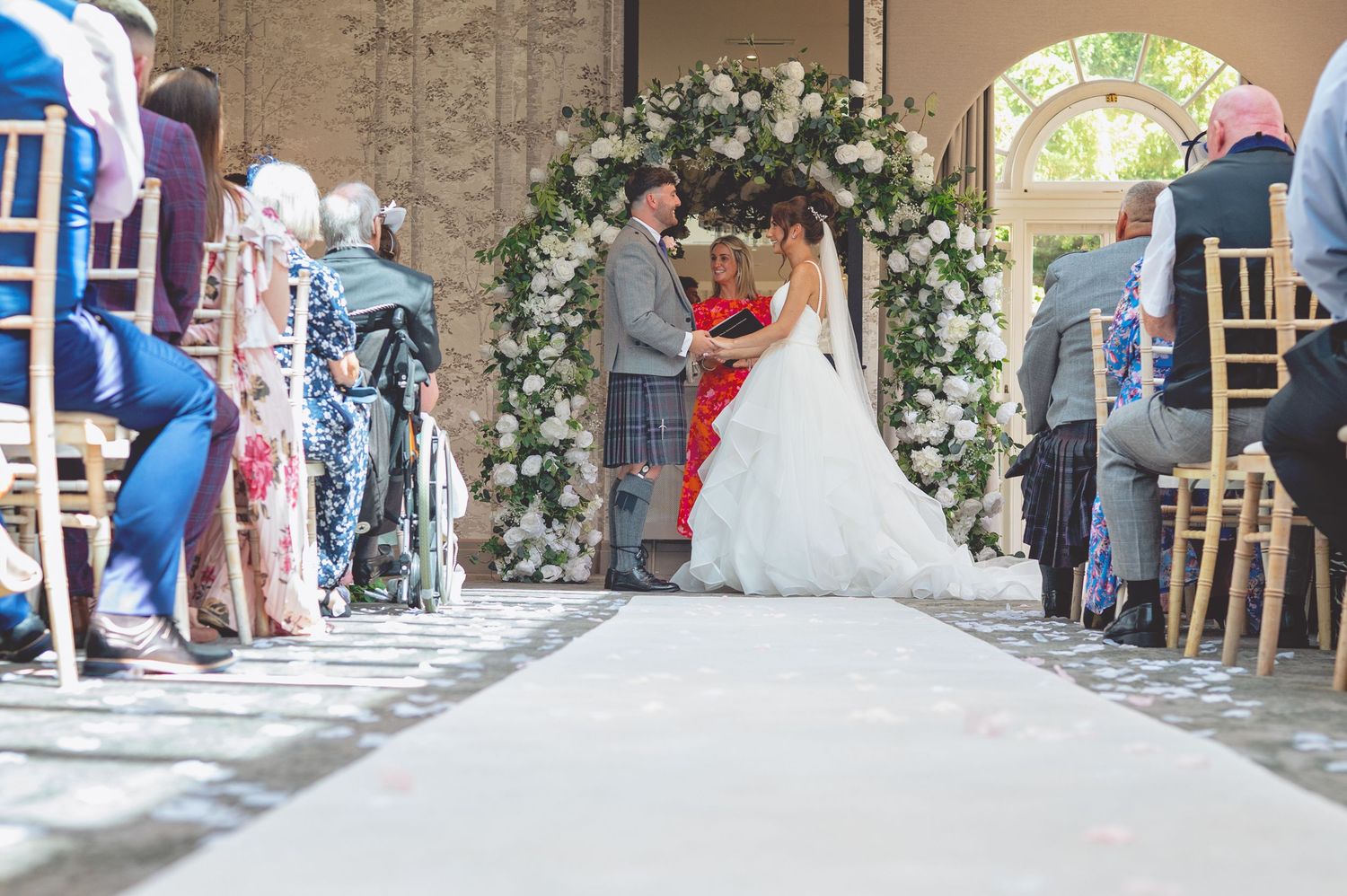 Wedding ceremony setup in an elegant indoor venue with floral arrangements and white chairs.