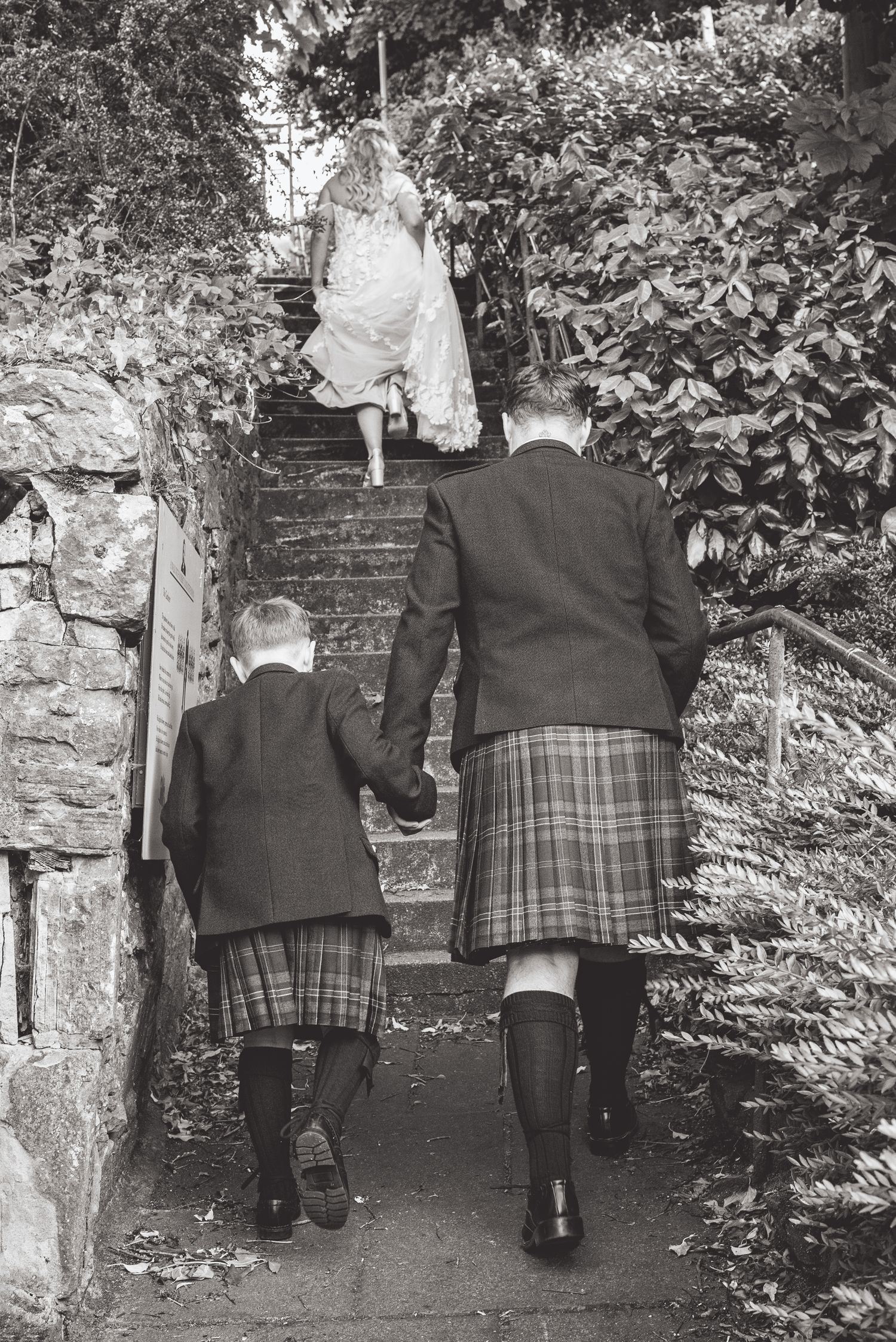 Wedding guests in formal attire and kilts walking along stone garden path.