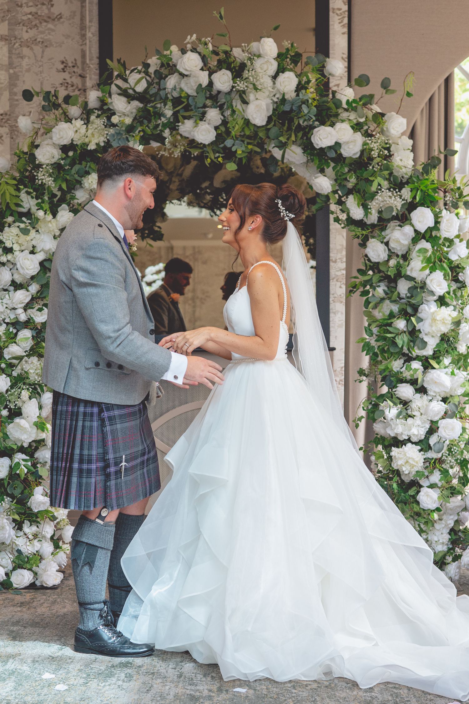 Wedding couple posing under an elegant white floral arch with roses and greenery.
