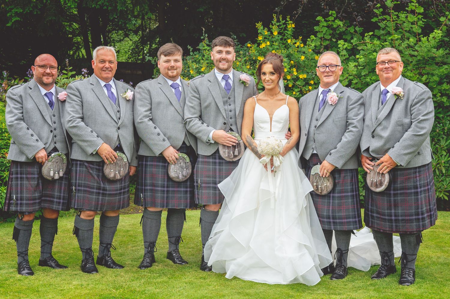 Wedding party members in grey suits and formal attire pose for group photo in garden.