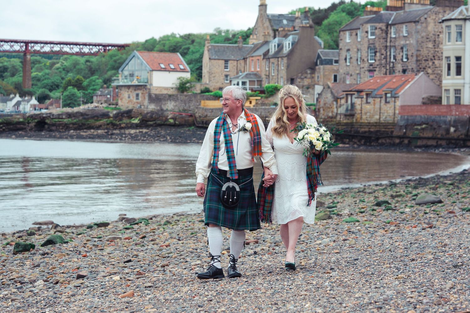 Bride and groom walking along a Scottish shoreline, man in tartan kilt, woman in white dress holding bouquet.