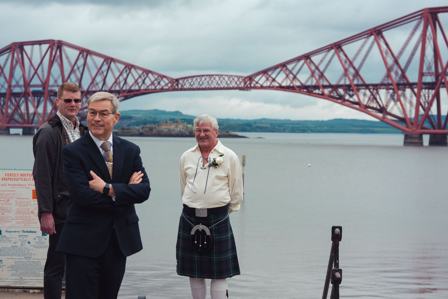 Three men posing near the Forth Rail Bridge in Scotland, one wearing a traditional kilt, with the red bridge over water behind.