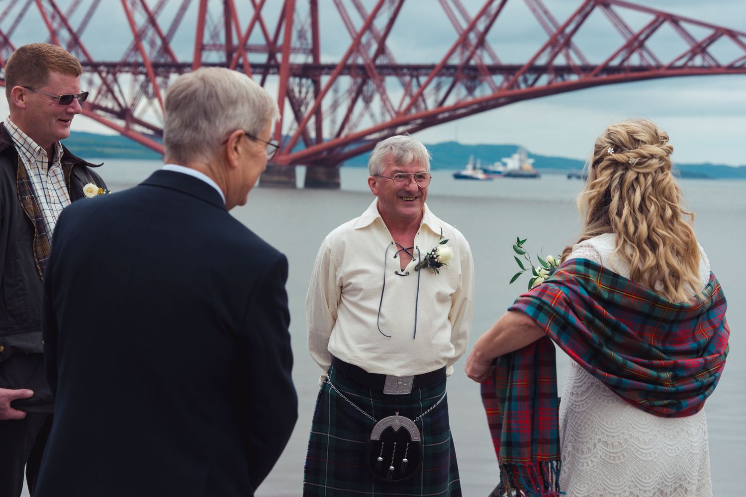 Wedding ceremony outdoors near the Forth Bridge in Scotland, groom in tartan kilt with guests and bride holding flowers.