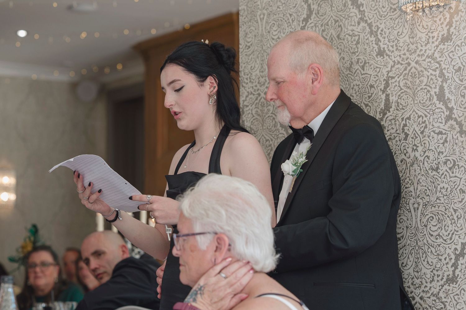Young woman in black dress reads speech beside elderly man in suit at a formal wedding reception.