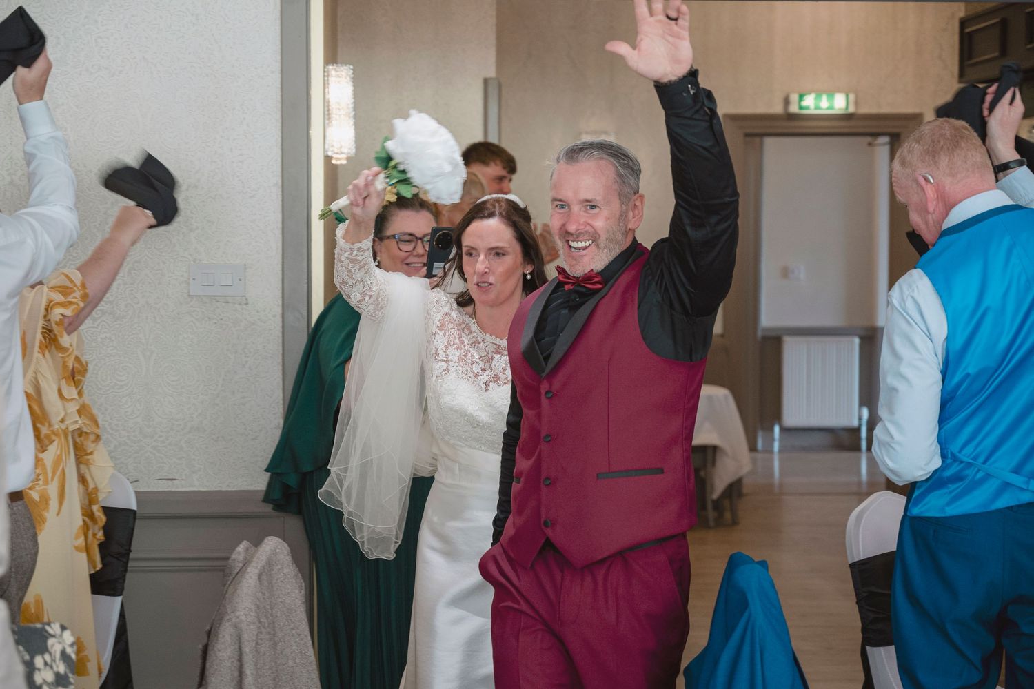 Happy couple celebrating their wedding reception, groom in burgundy suit raising hand, bride in white dress holding bouquet.
