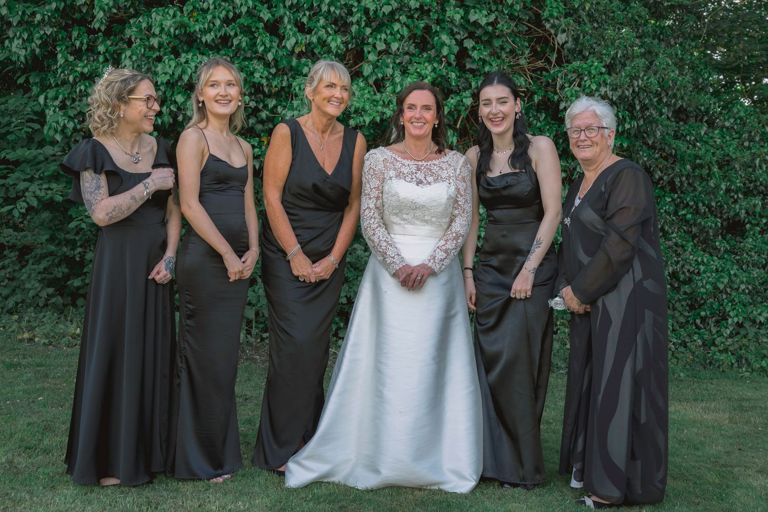 Bride in white lace gown poses with five bridesmaids in black dresses outdoors against a lush green ivy backdrop.