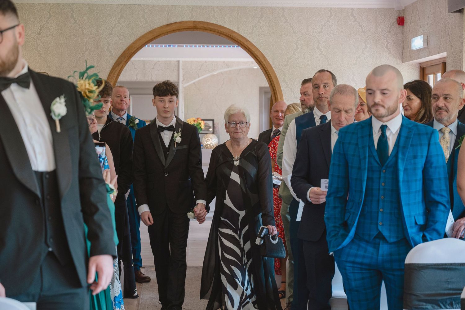 Wedding procession with guests walking through an arched doorway in a formal venue, wearing elegant attire including tuxedos and suits.