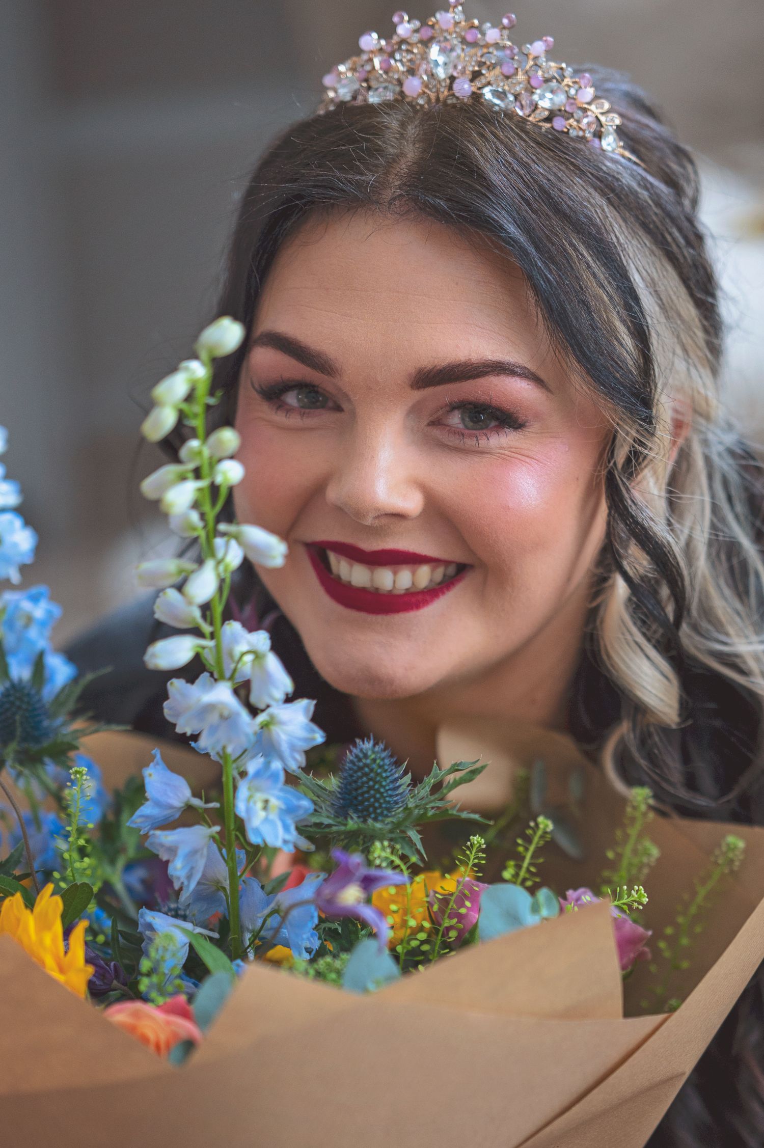 Smiling bride wearing floral tiara holding colorful wedding bouquet with blue and pink flowers.