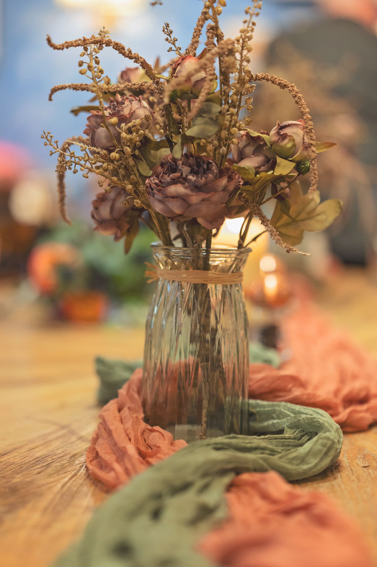 Rustic floral centerpiece with dusty pink roses in a glass vase on a wooden table with coral and sage green fabric runners.