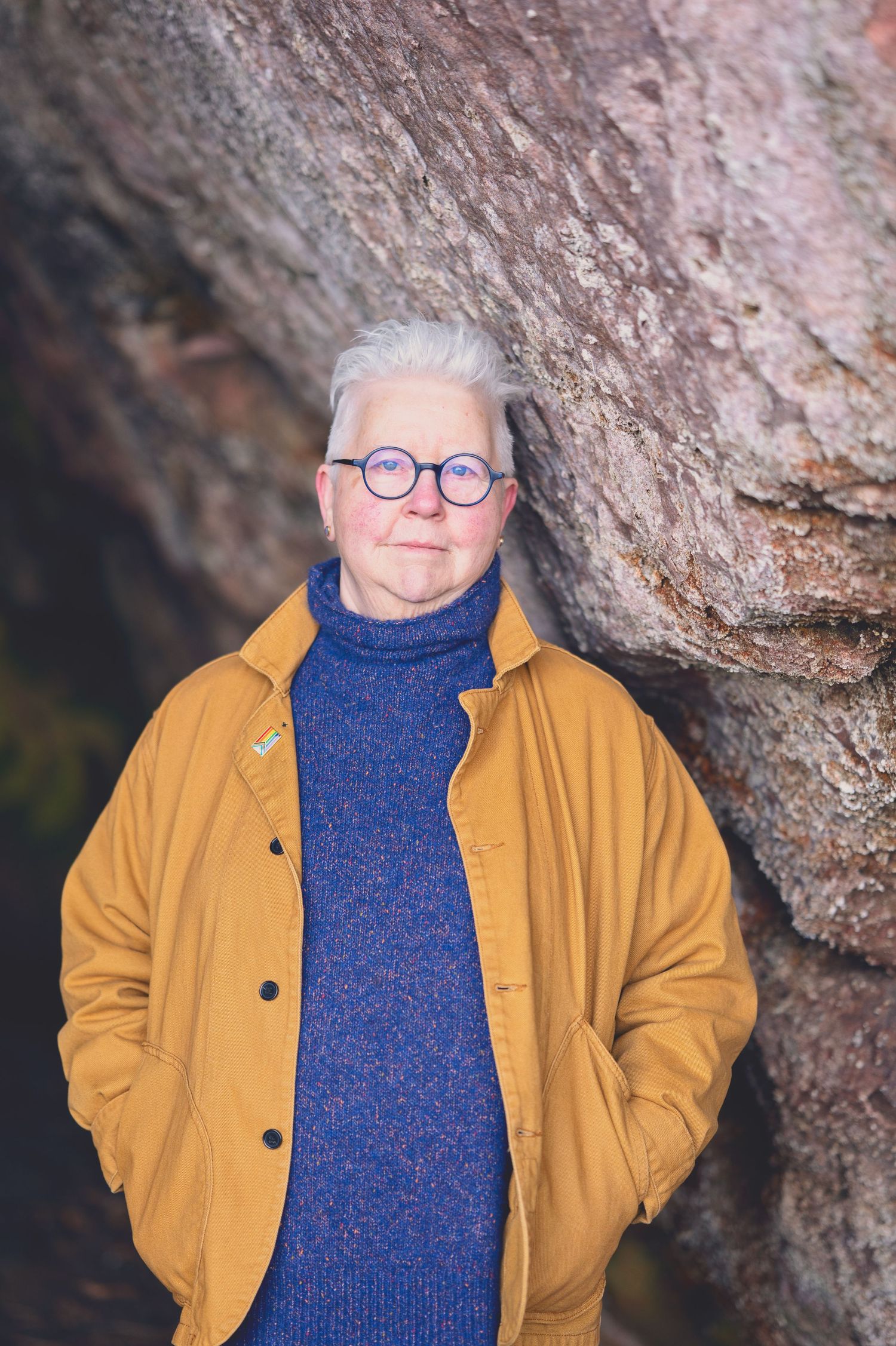 The Author Val Mcdermid with short white hair and round glasses wearing a mustard jacket and blue turtleneck sweater in the Weymss caves