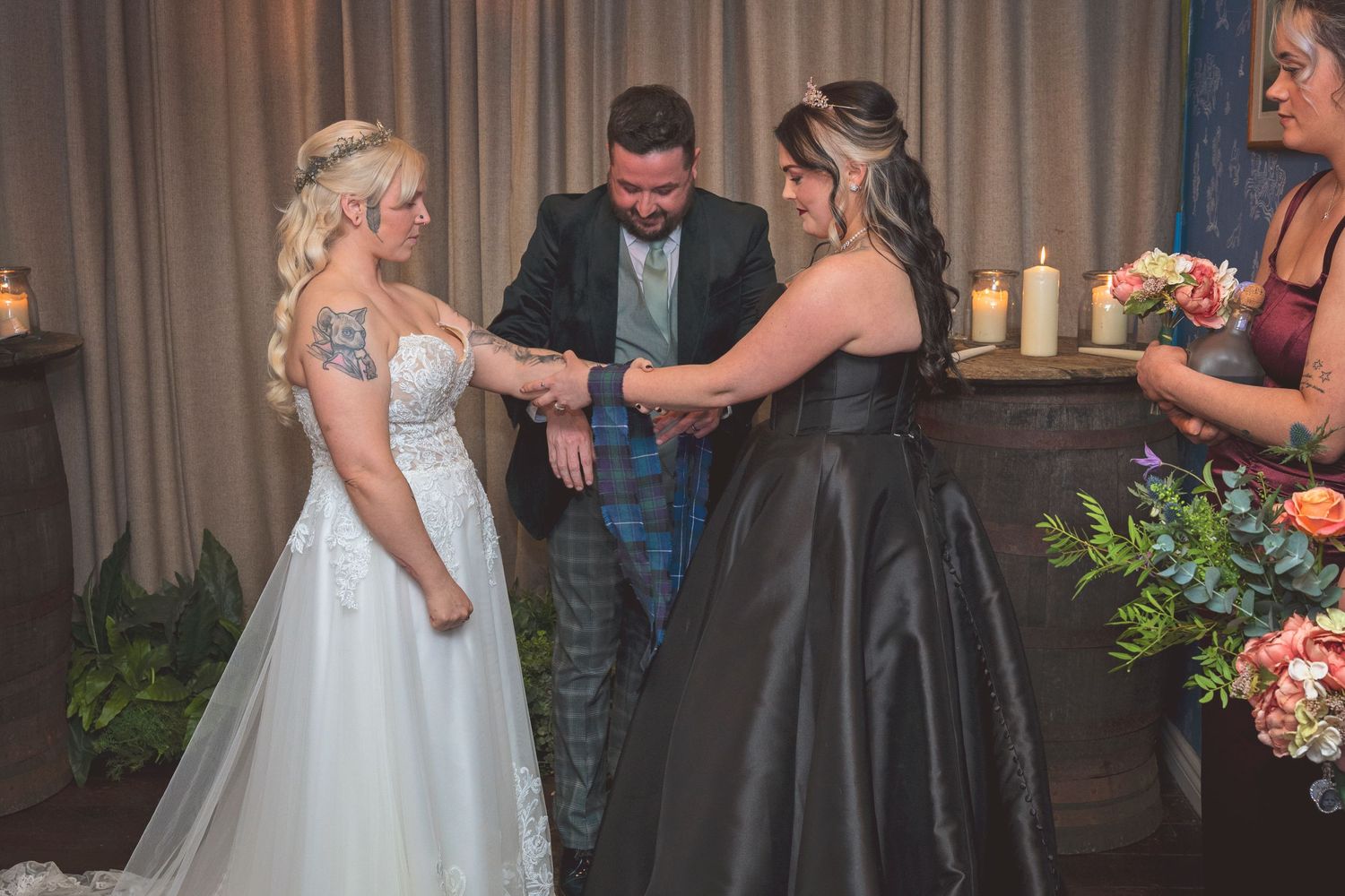 Groom in tartan kilt performs hand-fasting ceremony with two brides in wedding attire at candlelit altar.