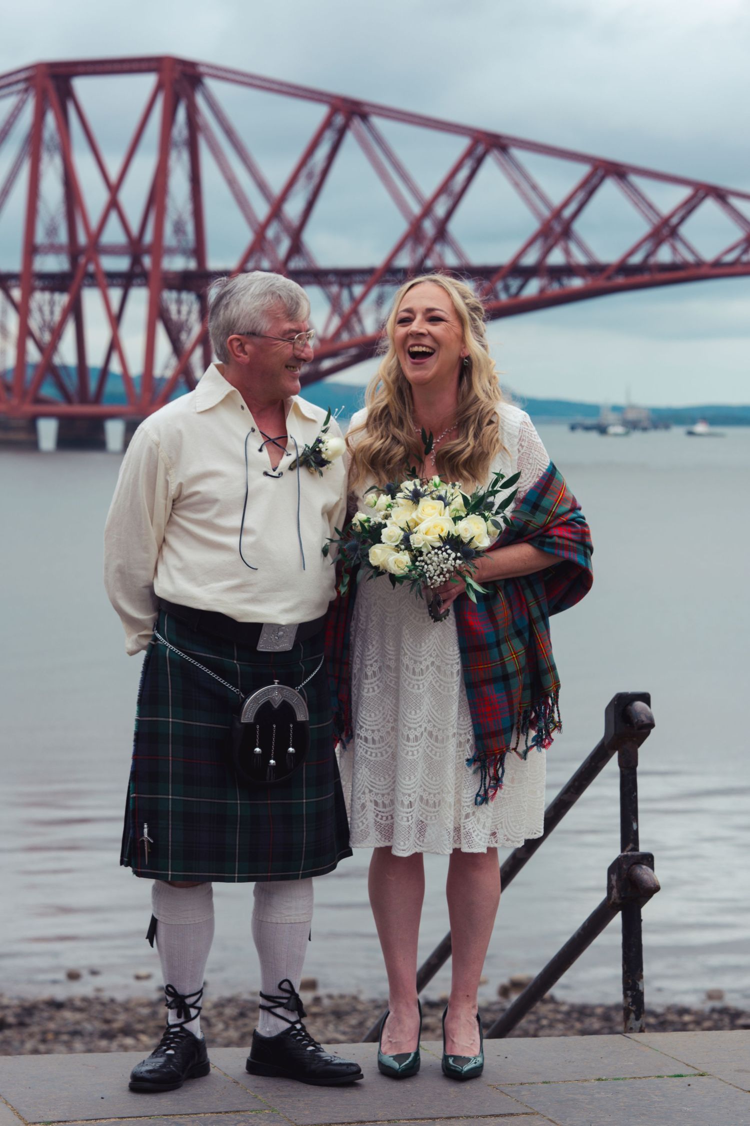Couple laughing in Scottish wedding attire by the Forth Bridge, man in kilt, woman in white dress with tartan shawl.