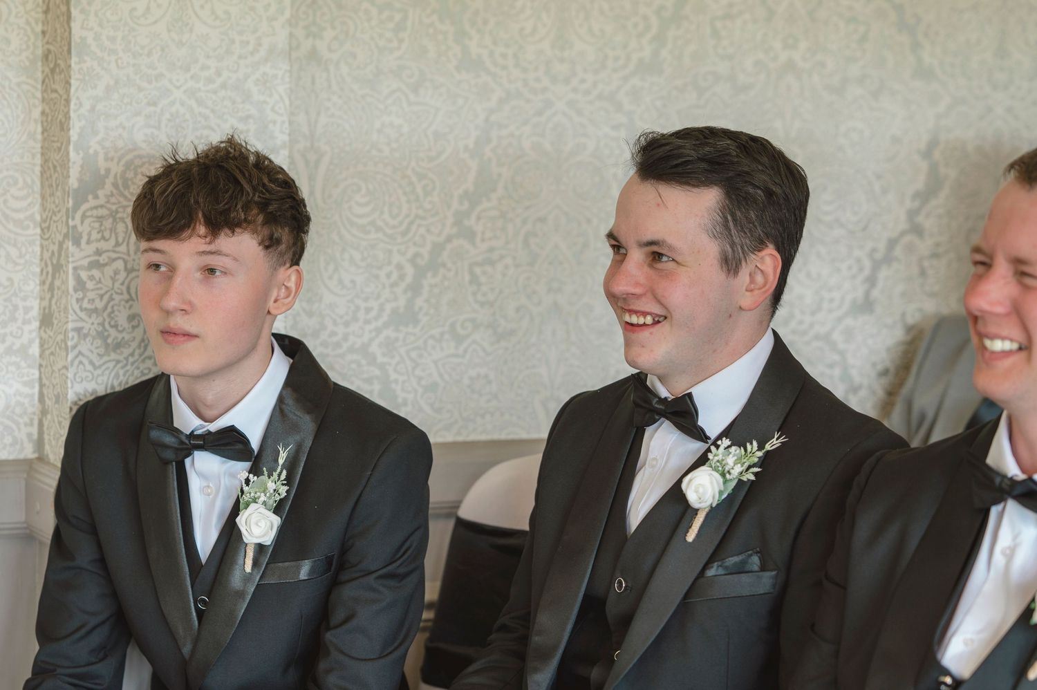 Three young men in black tuxedos with white boutonnieres seated at a formal wedding ceremony.