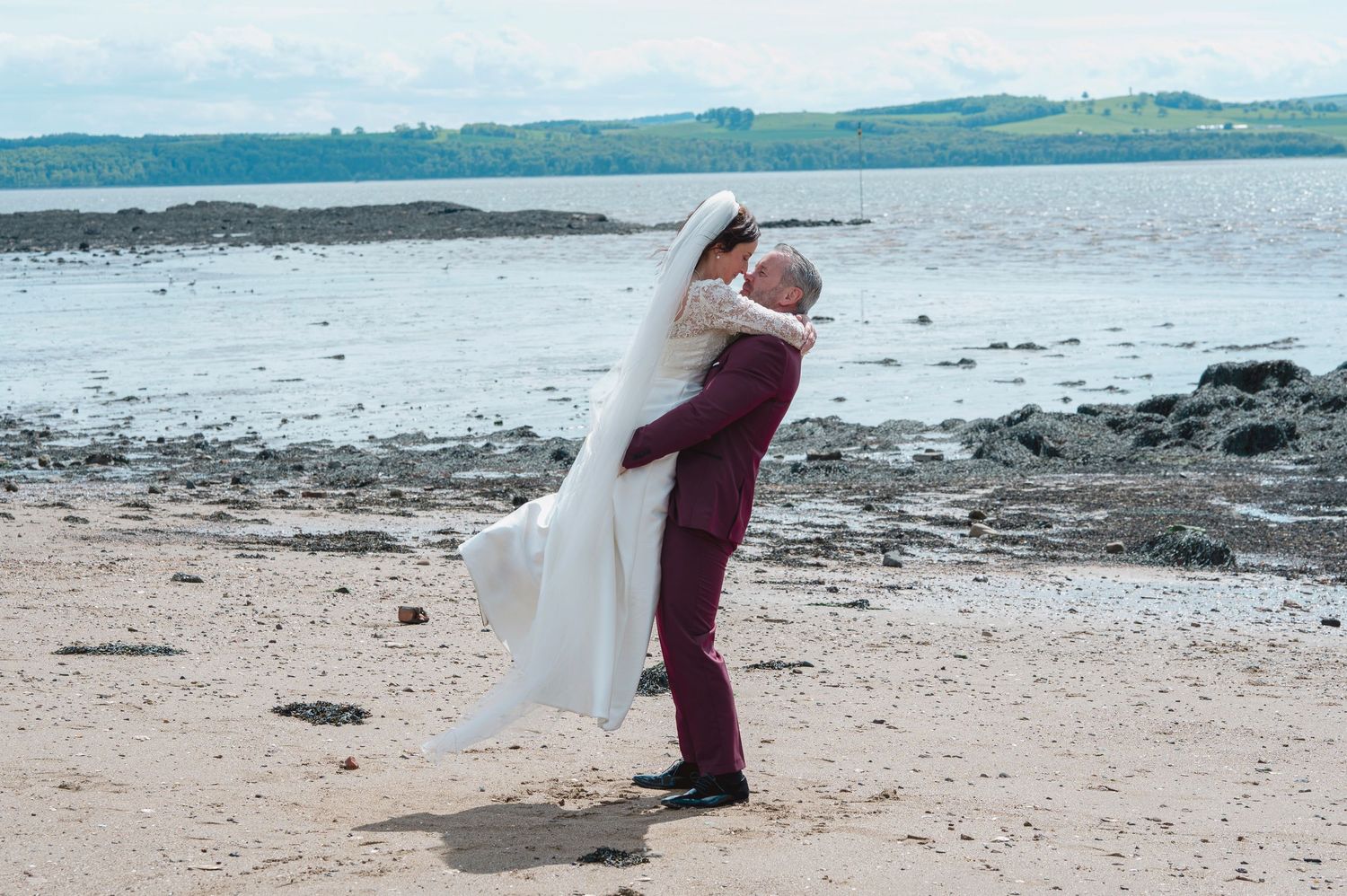 Groom in burgundy suit lifts bride in white wedding dress on sandy beach with calm sea backdrop.