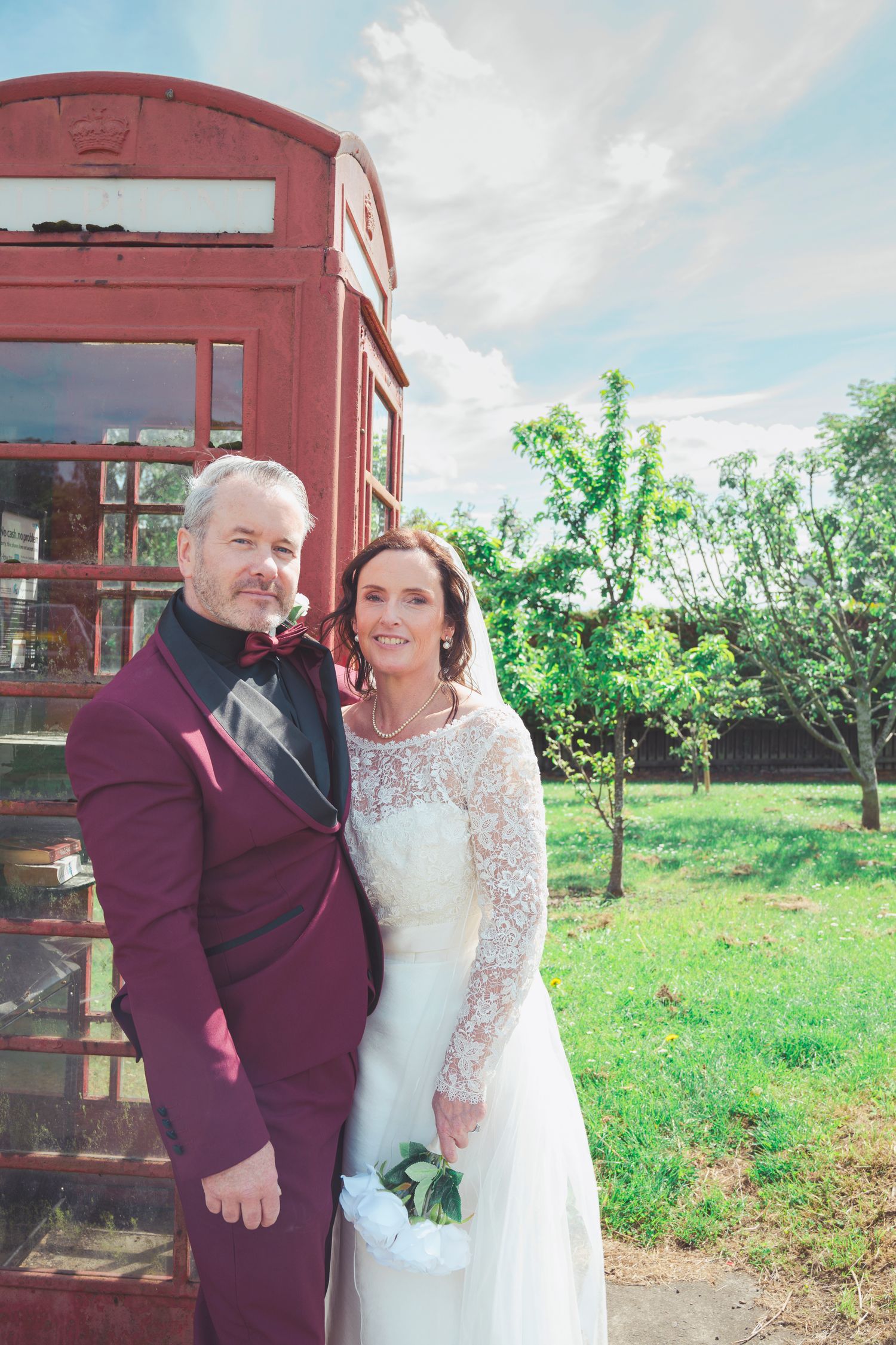 Bride and groom pose in front of a red British phone box on their wedding day, surrounded by lush greenery.