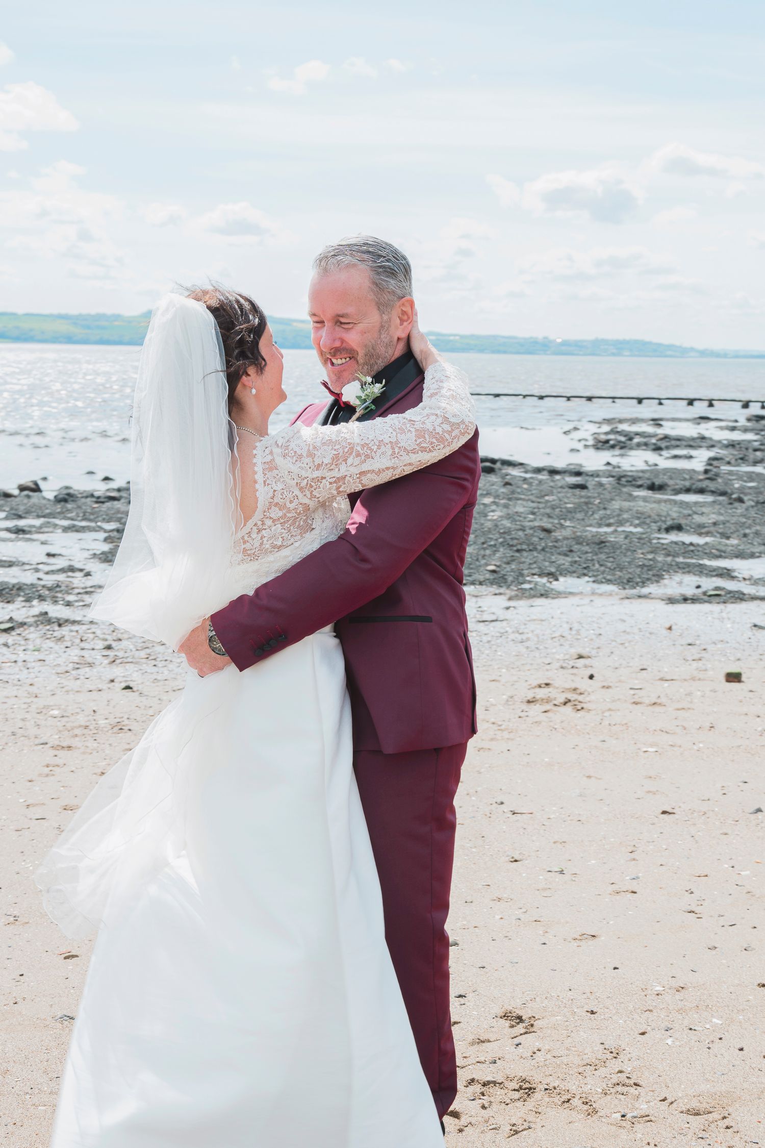 A bride and groom embrace on a sandy beach, with the groom in a burgundy suit and bride in a white gown with veil.