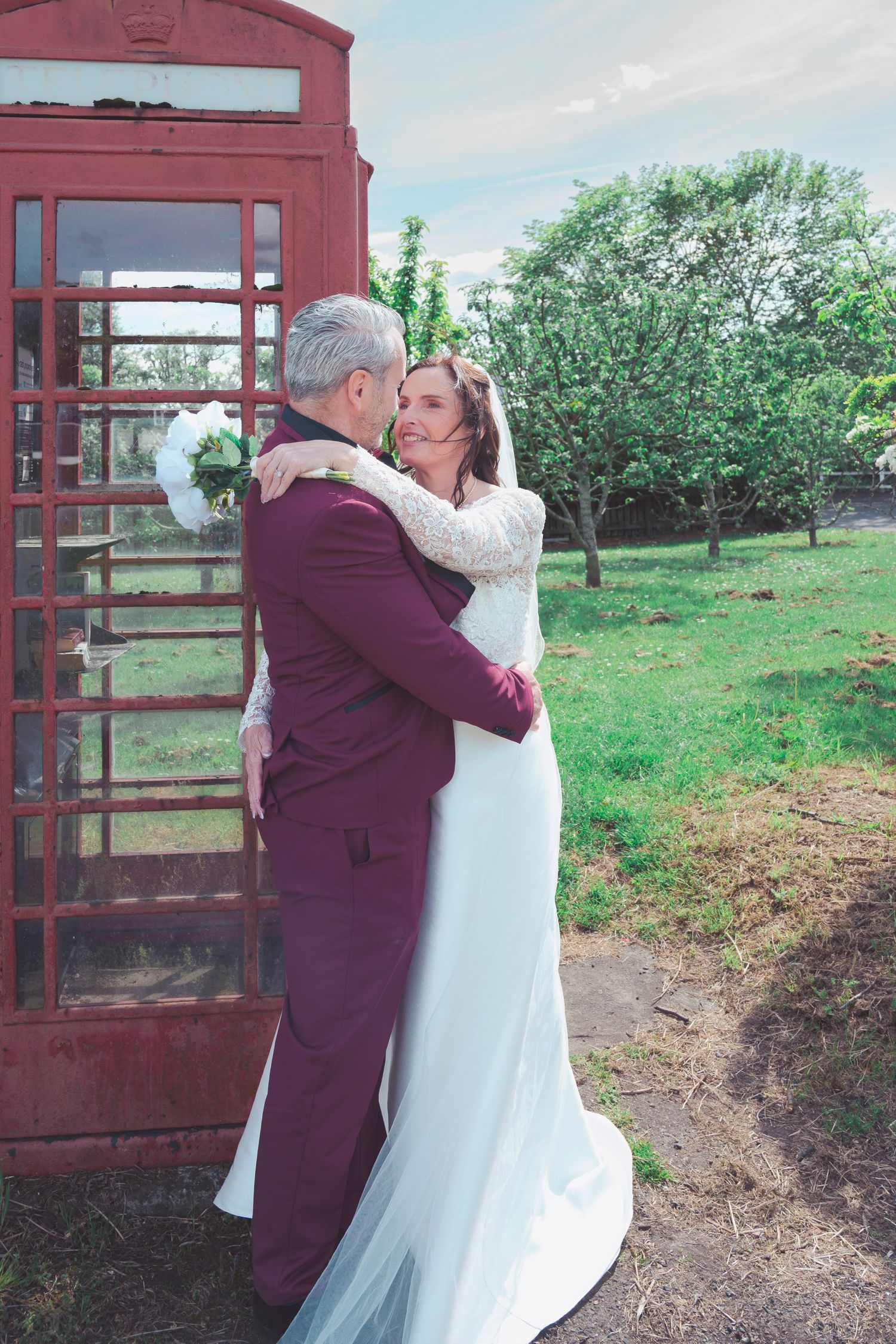 Bride and groom embracing and kissing beside a red British phone box on their wedding day.