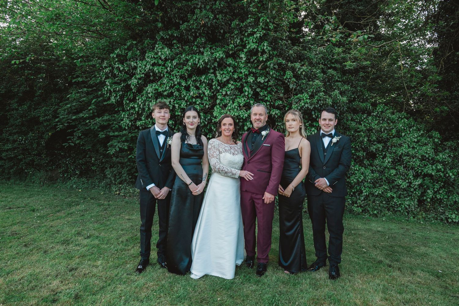 Formal family group photo with six people in elegant attire posing outdoors against lush green foliage.