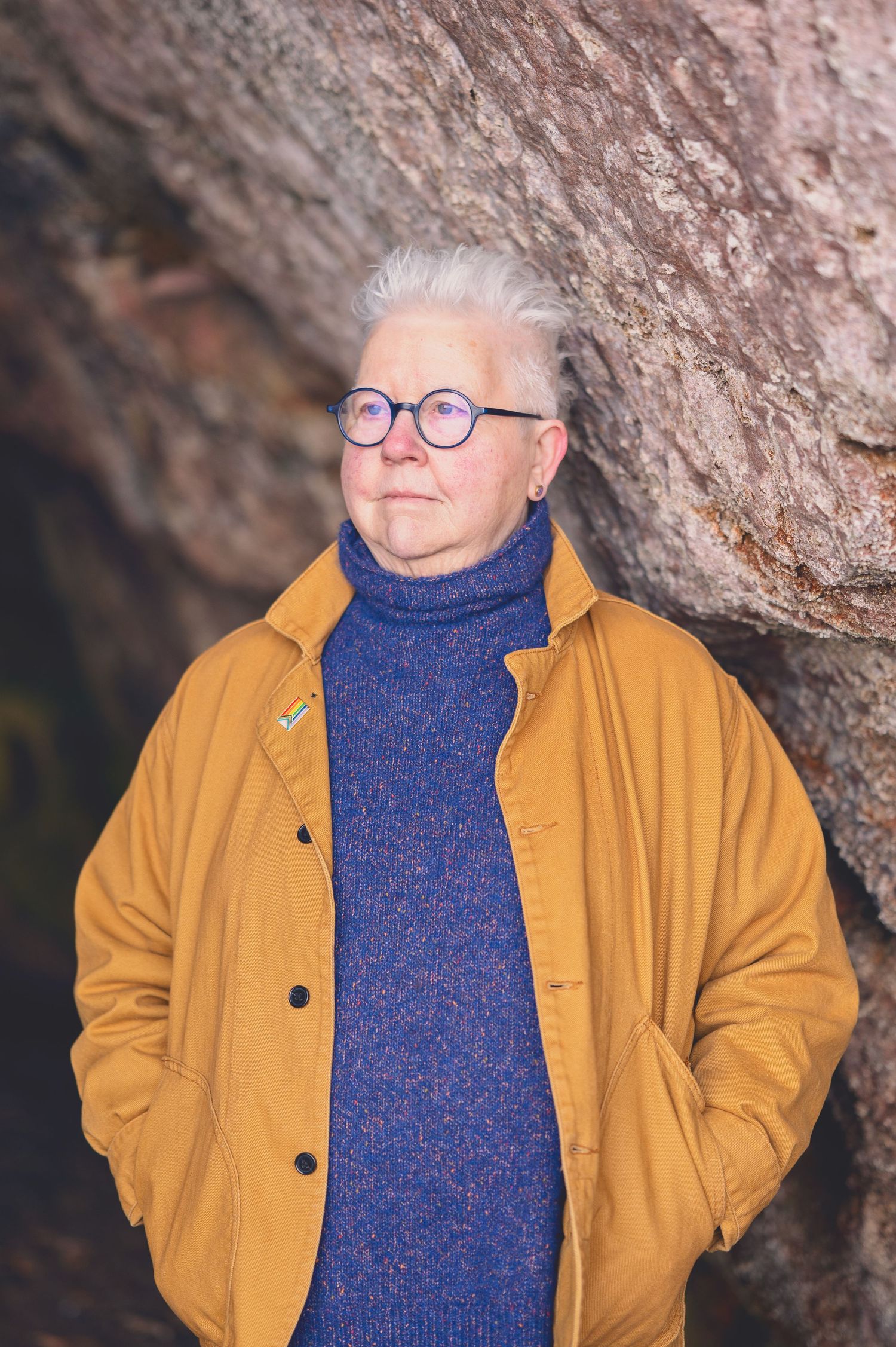 The Author Val Mcdermid with short white hair and round glasses wearing a mustard jacket and blue turtleneck sweater in the Weymss caves