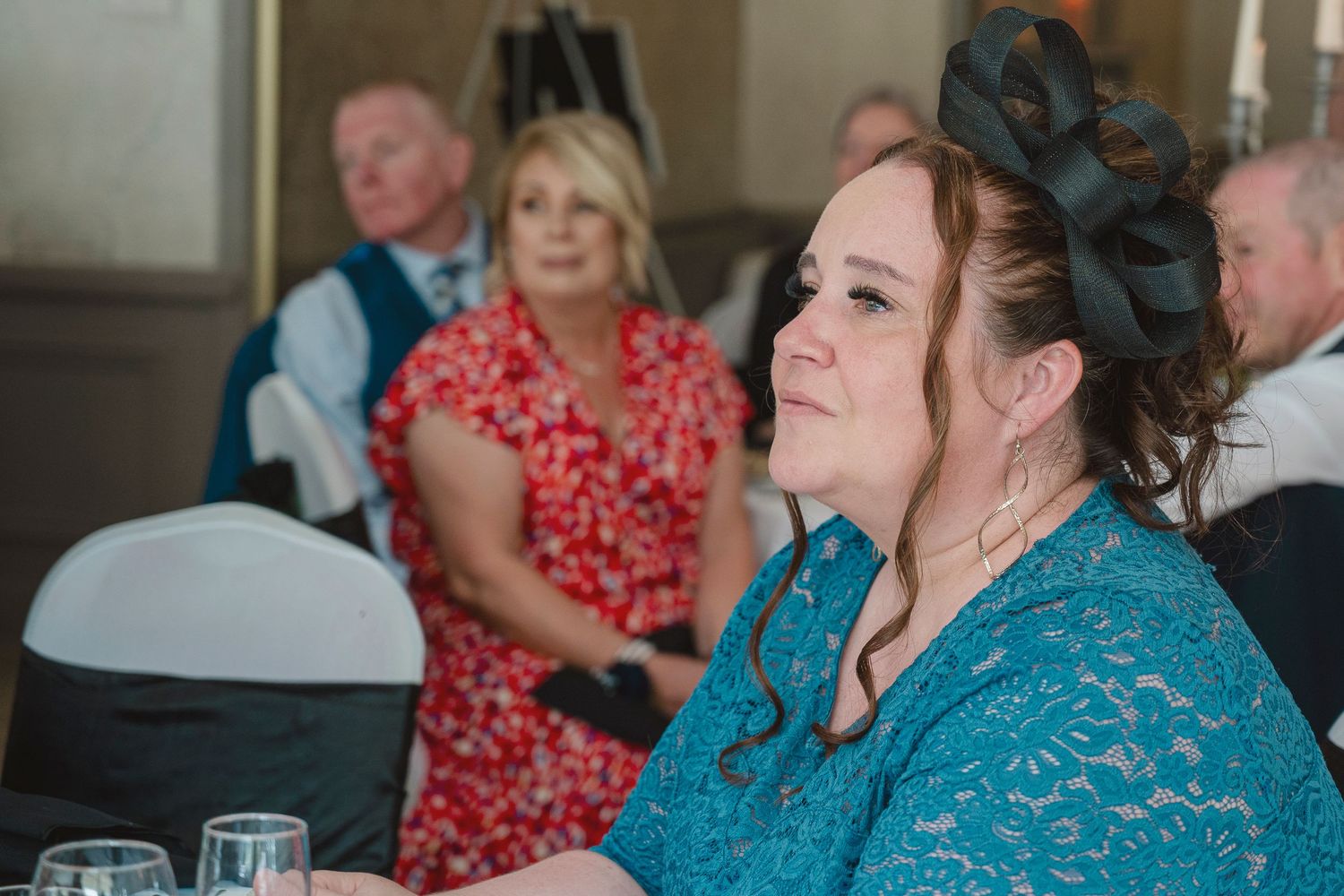 Woman in teal lace dress and black fascinator seated at wedding reception table, guests in background.