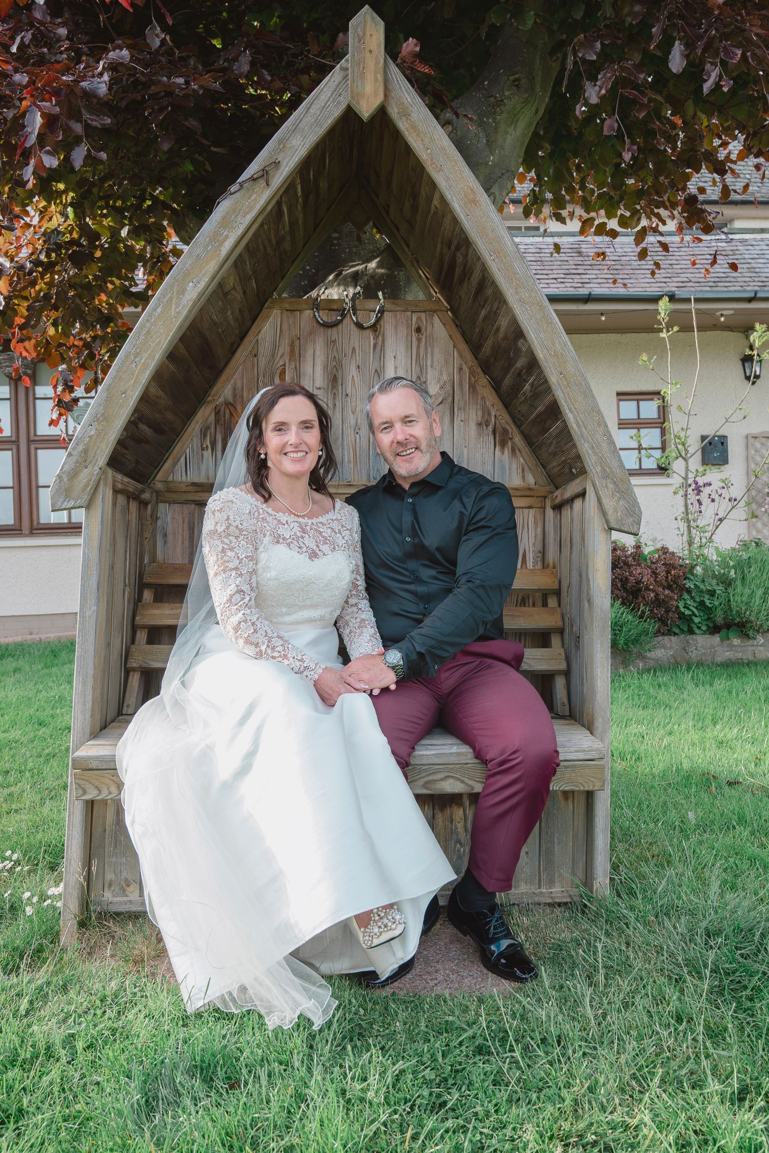 A bride and groom sit together in a wooden arch garden seat, smiling, surrounded by lush greenery on their wedding day.