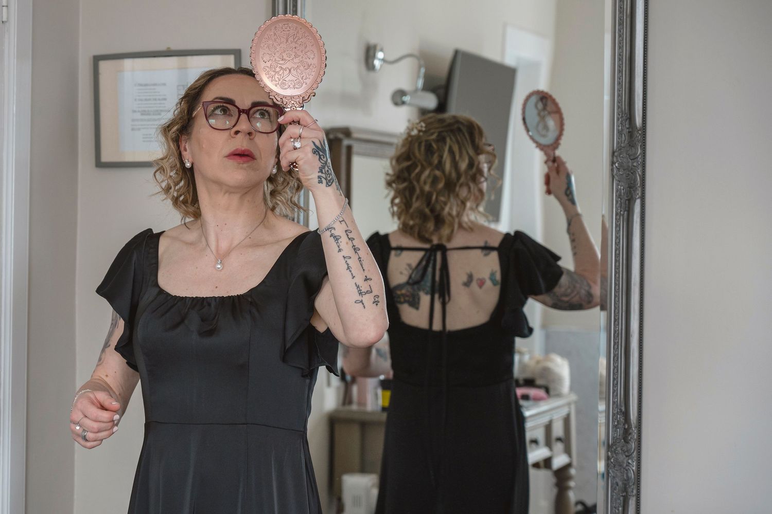 Tattooed woman in black dress holds pink mirror, reflected in large floor mirror in bright room.