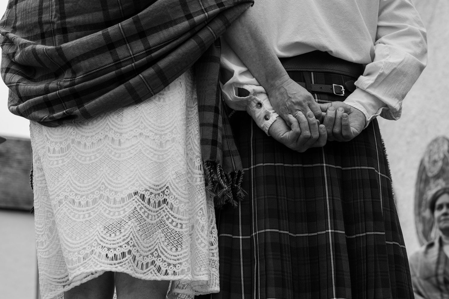 Black and white photo of a couple in Scottish attire holding hands, wearing a kilt and lace skirt.