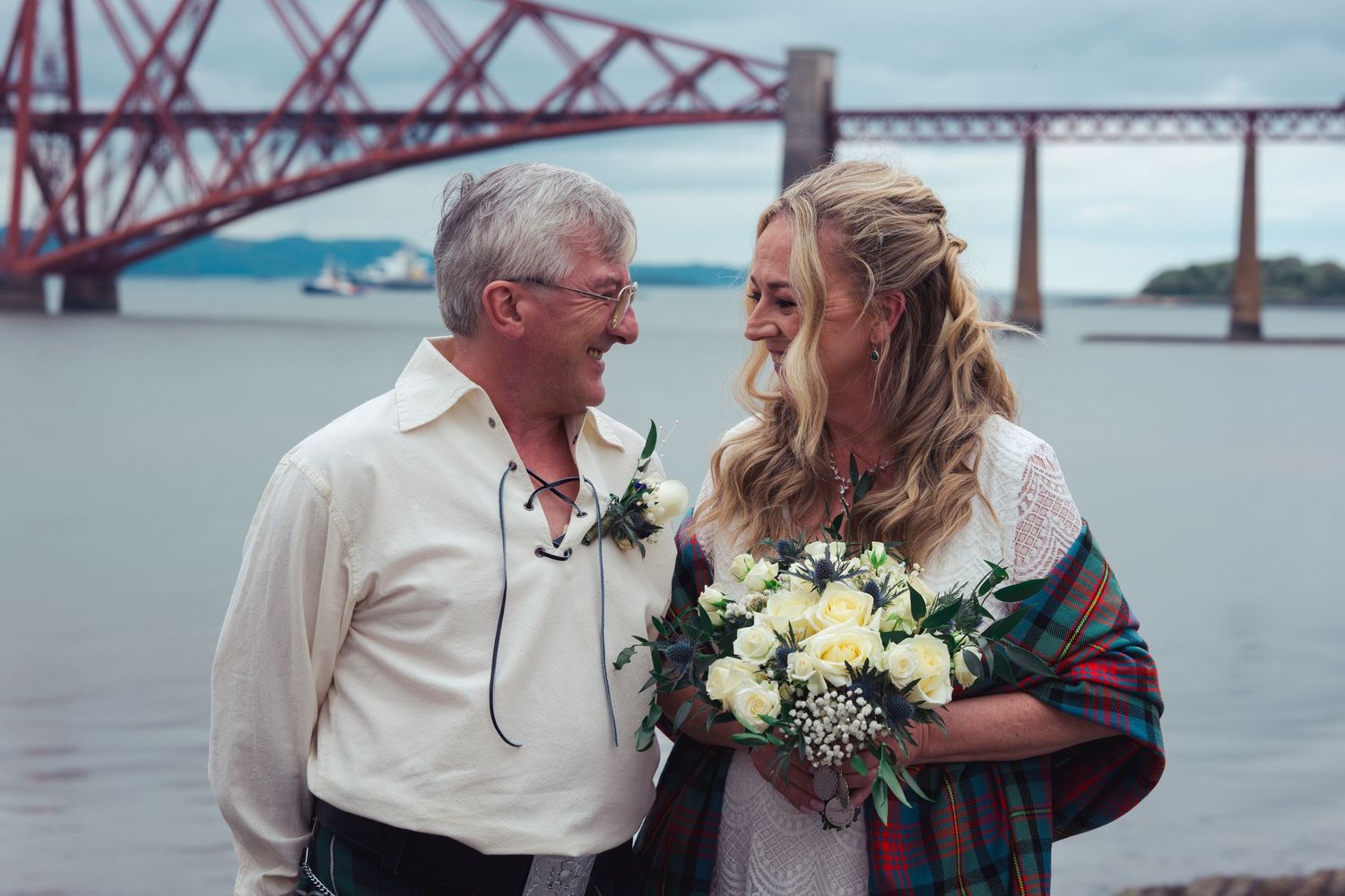 Bride and groom smiling at each other near the Forth Bridge in Scotland, bride holding white flower bouquet.