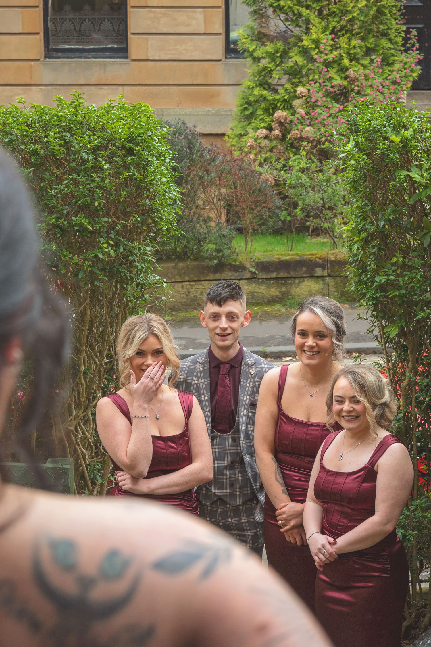 Bridesmaids in burgundy dresses and groomsman in plaid suit stand in garden courtyard during outdoor wedding ceremony.