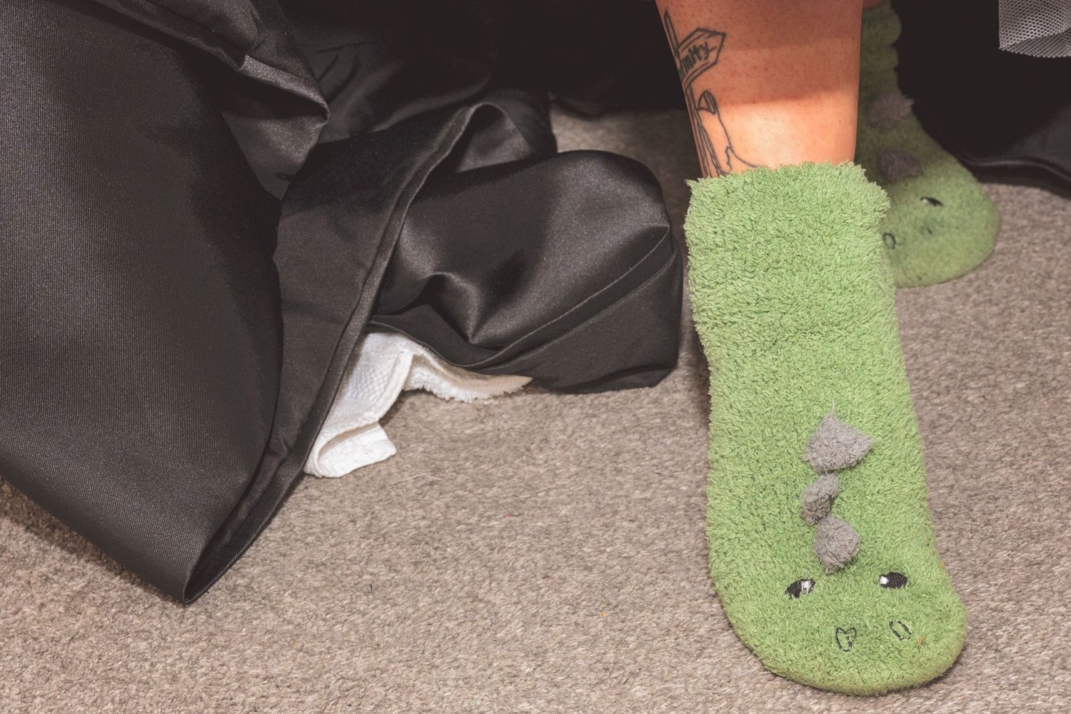 Close-up of green fuzzy dinosaur novelty socks worn near black bags on carpet floor.