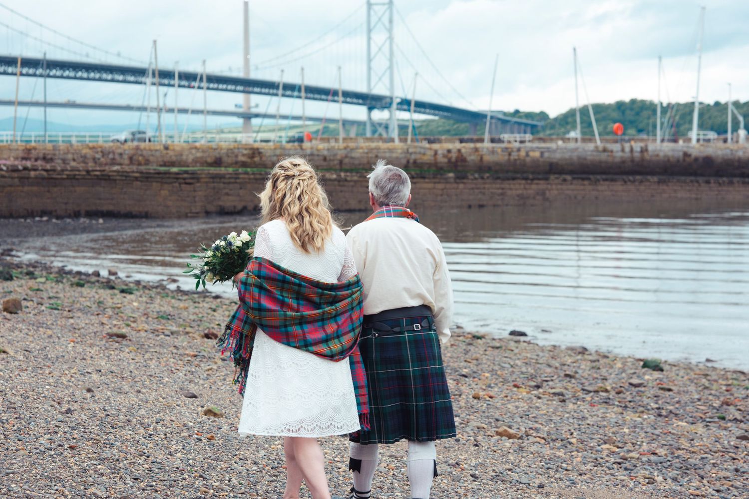 Couple in Scottish tartan attire walking along a pebble beach with a suspension bridge in the background.