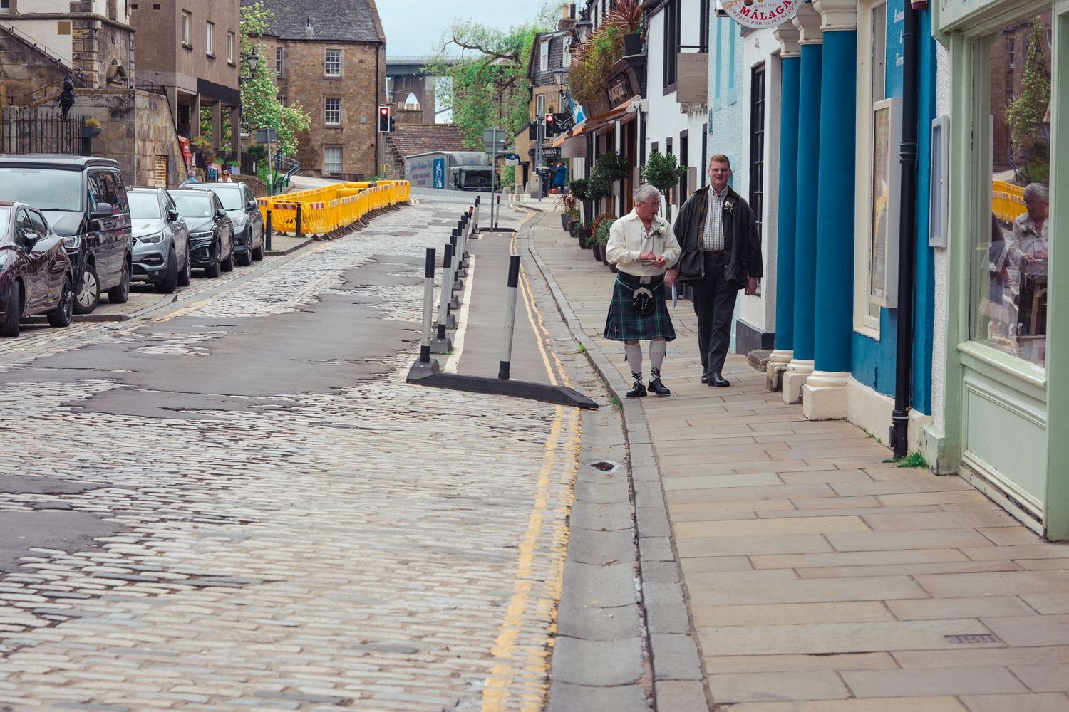 Man in traditional Scottish kilt walking on a historic cobblestone street lined with colorful shops in Edinburgh.