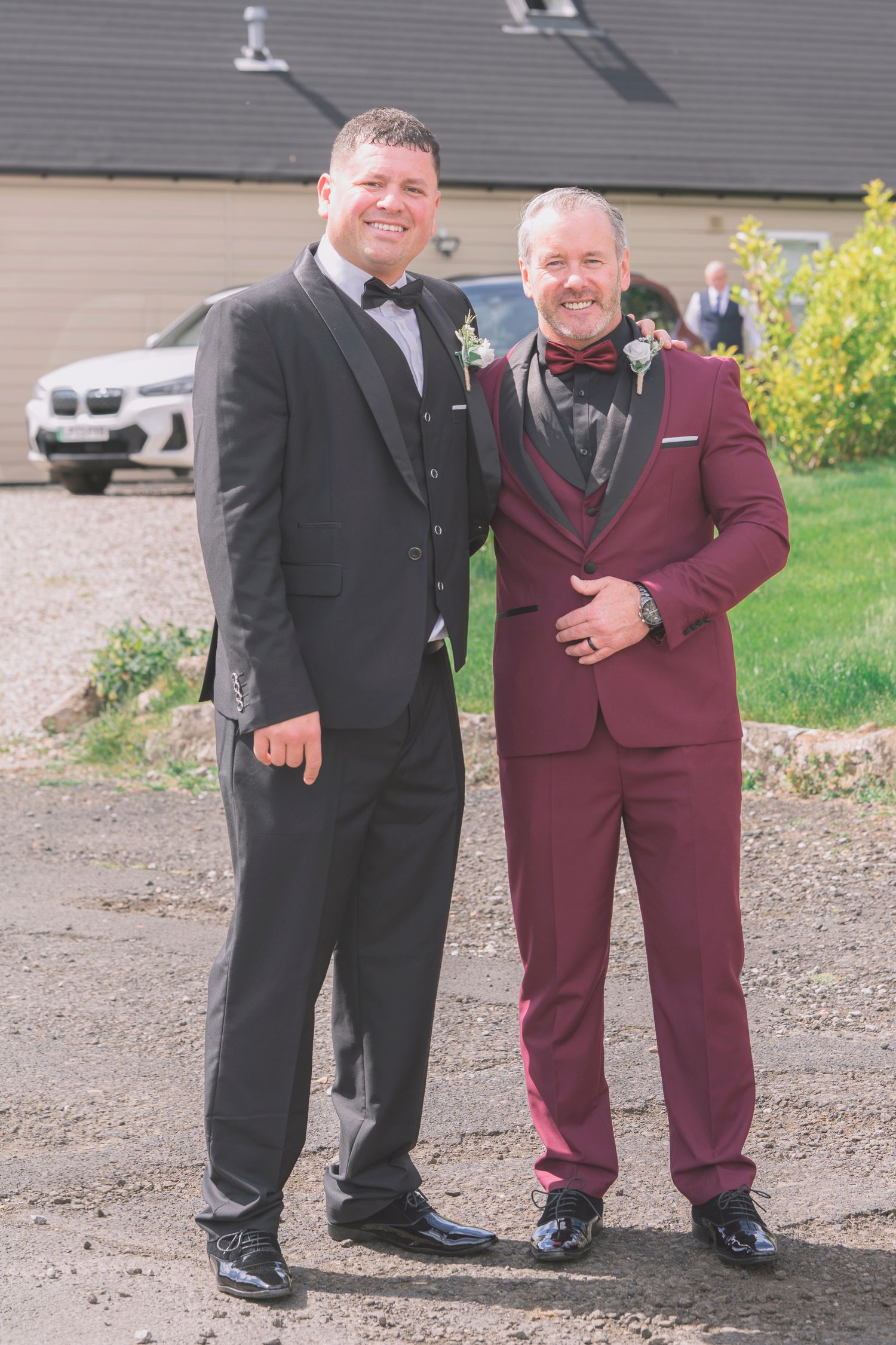 Two men in formal suits with boutonnieres posing outdoors at a wedding venue on a sunny day.
