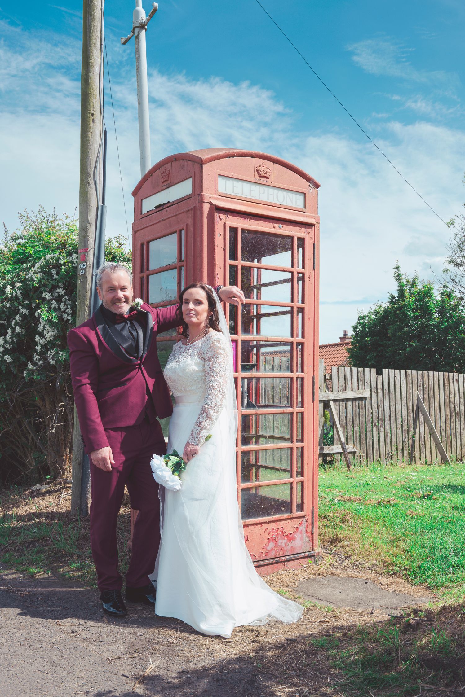 Bride and groom posing beside a classic red British phone box on their wedding day in a sunny outdoor setting.