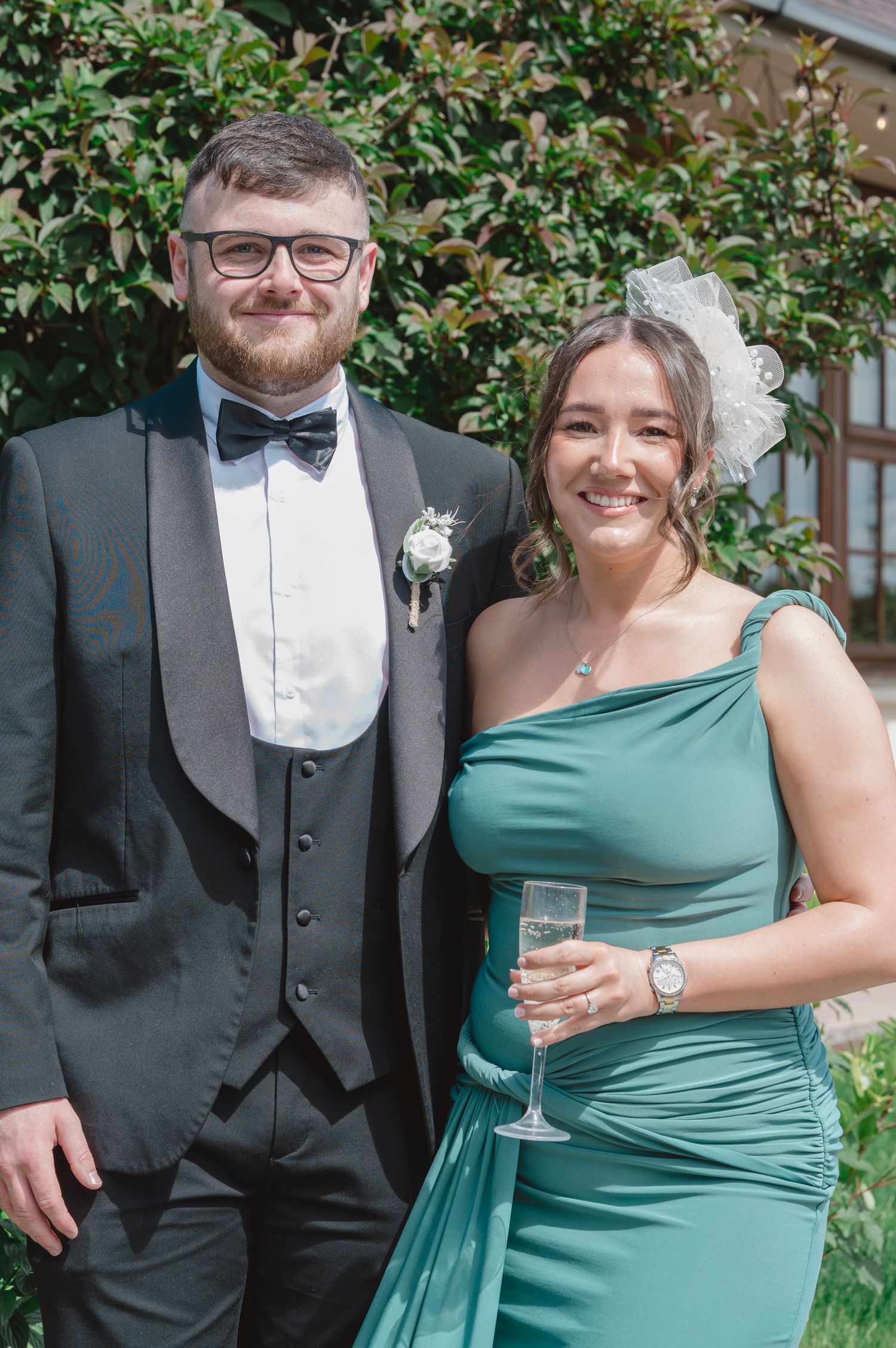 Couple dressed in formal attire at a wedding, man in tuxedo and woman in teal gown holding champagne.