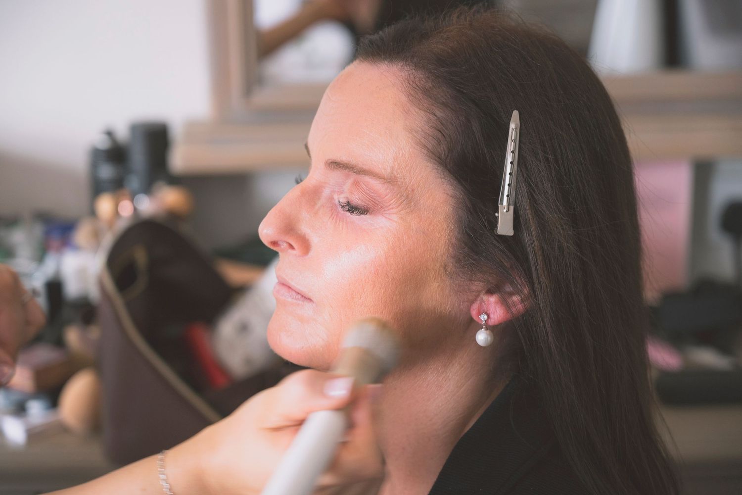 A woman getting professional makeup applied with a brush, wearing a pearl earring and hair clip.
