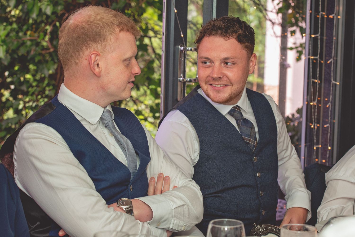Two wedding guests in formal waistcoats share a moment at the reception.