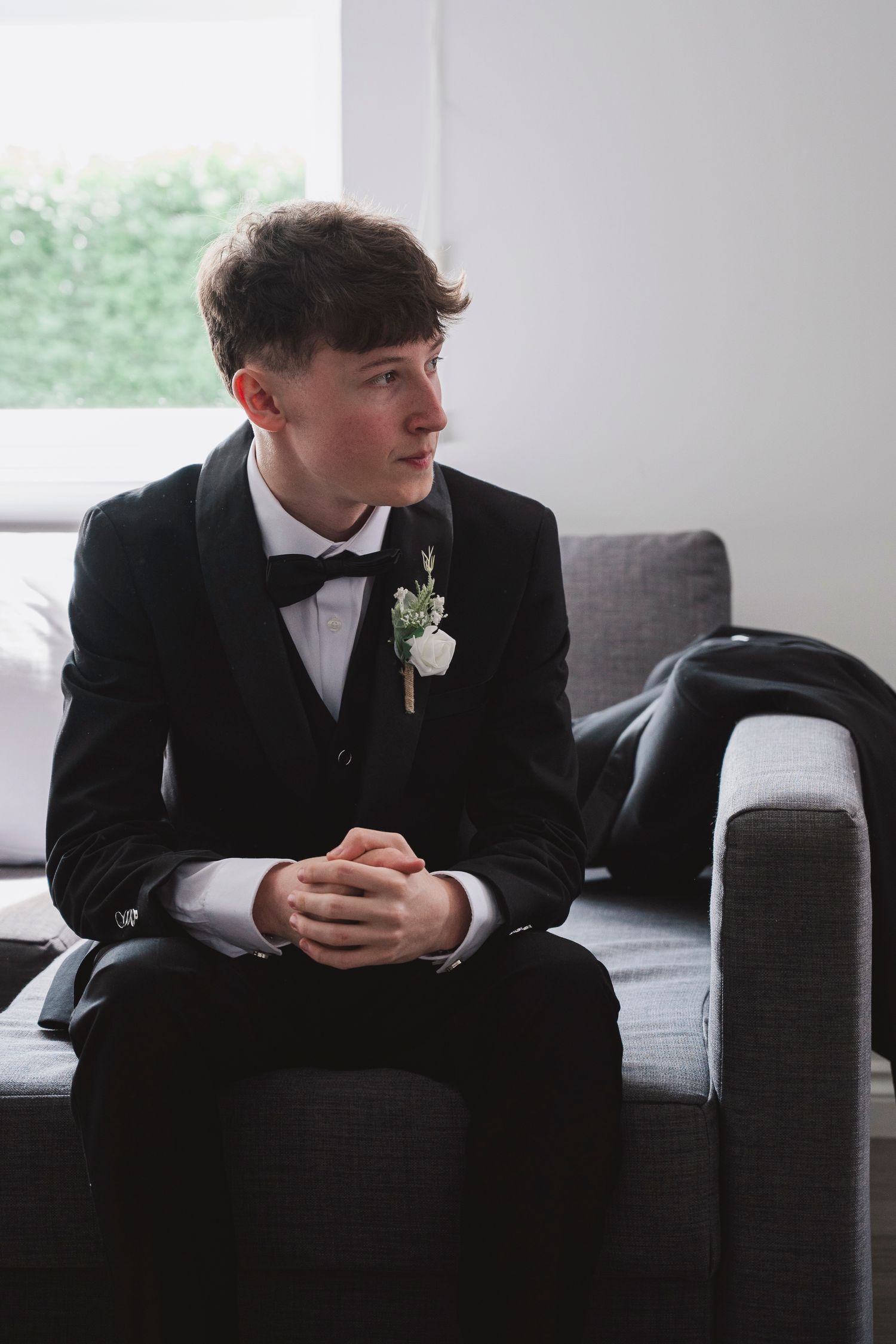 Young groom in black tuxedo with boutonniere sits on gray sofa, hands clasped, looking away thoughtfully.