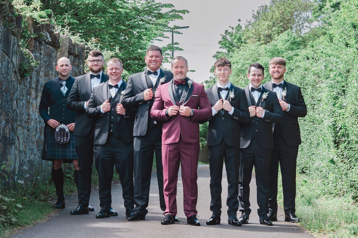 Groom in burgundy suit stands with groomsmen in black tuxedos and one in a kilt on a tree-lined path.